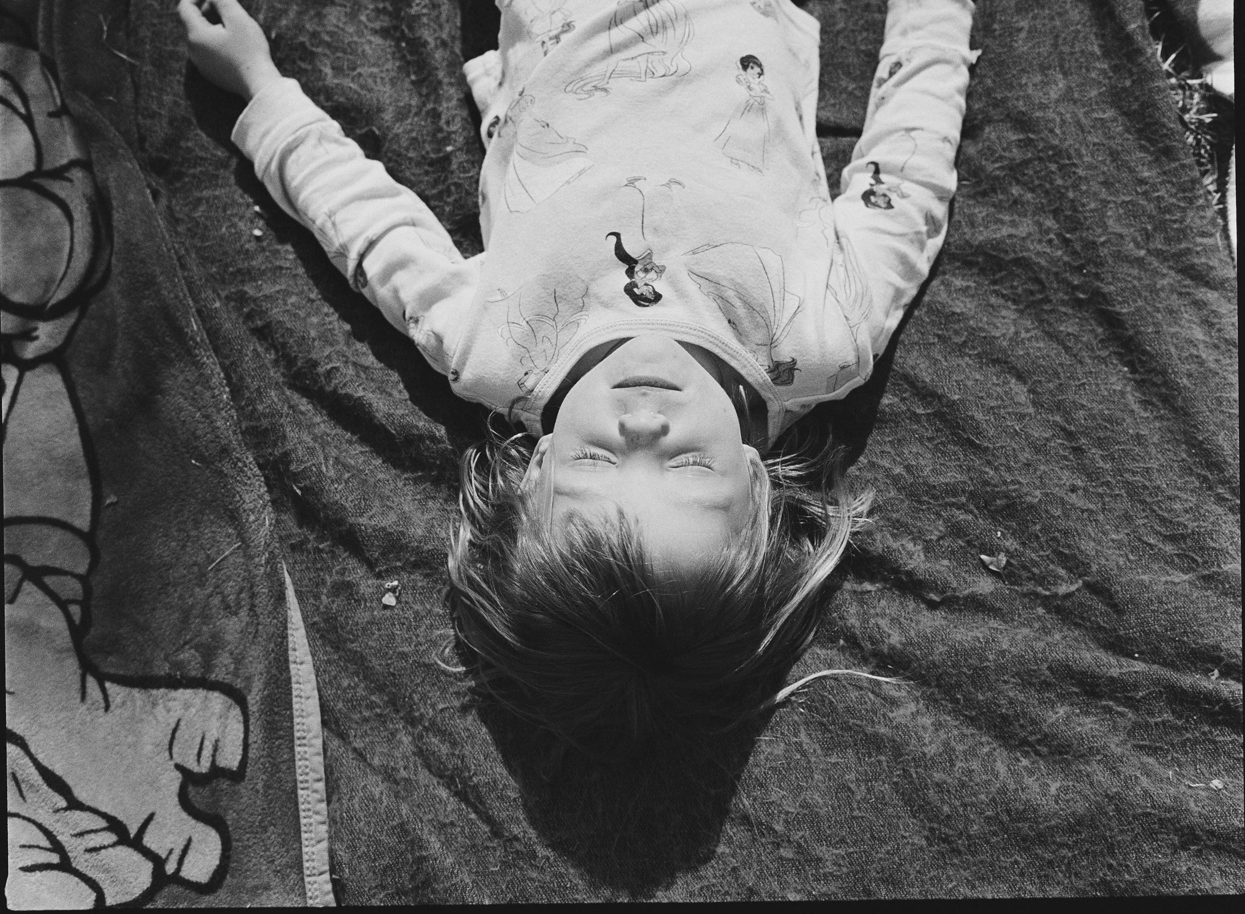 A young girl is lying on her back on a textured surface, possibly a blanket or rug, with her eyes closed and head tilted slightly to one side. She is wearing a long-sleeved shirt with cartoon princess characters on it. Her hair is down, spread over t