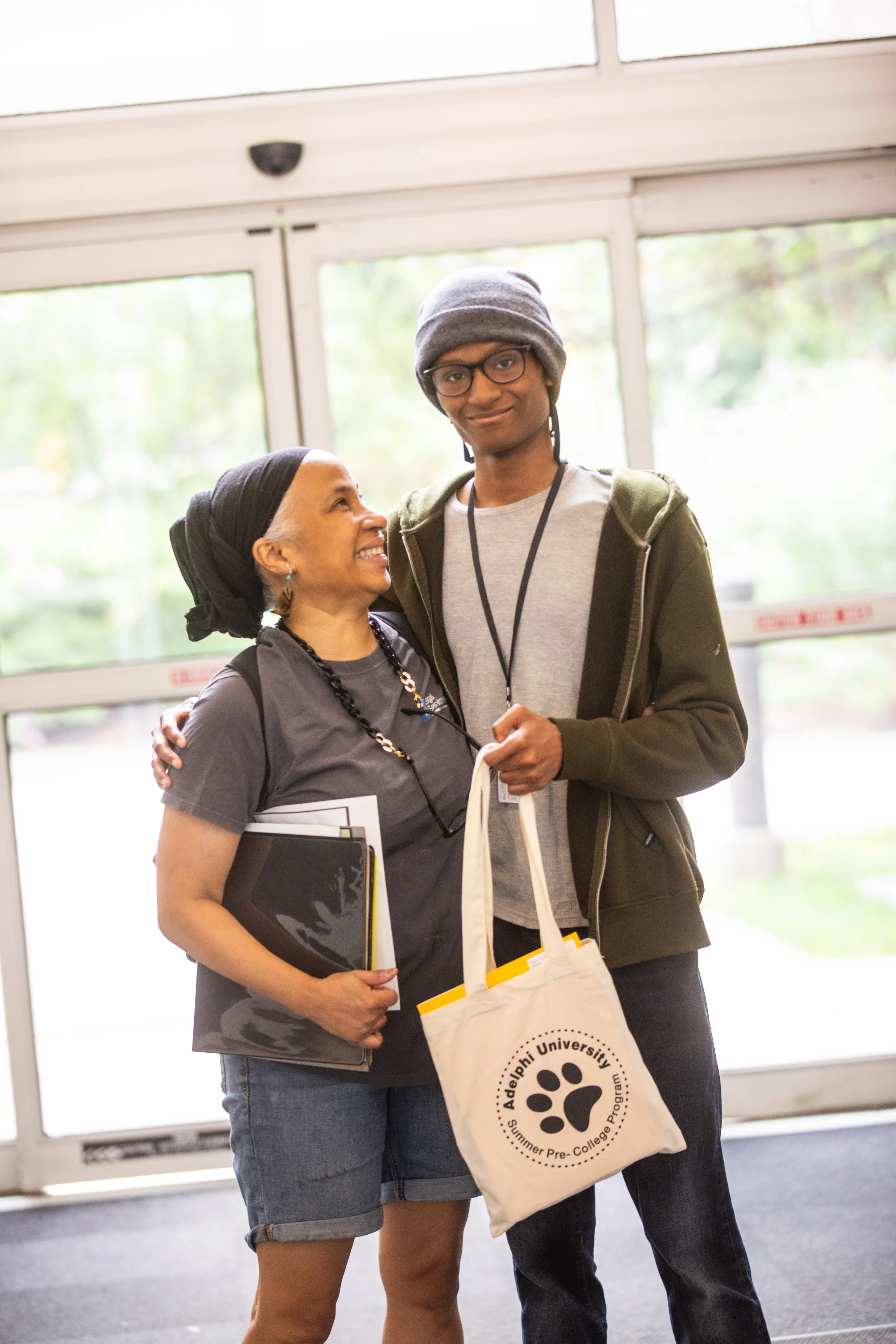 A woman and young man standing together indoors, smiling and embracing. The woman holds folders, and the young man holds a tote bag with 'Adelphi University Summer Pre-College Program' printed on it. Background shows glass doors and greenery outside.