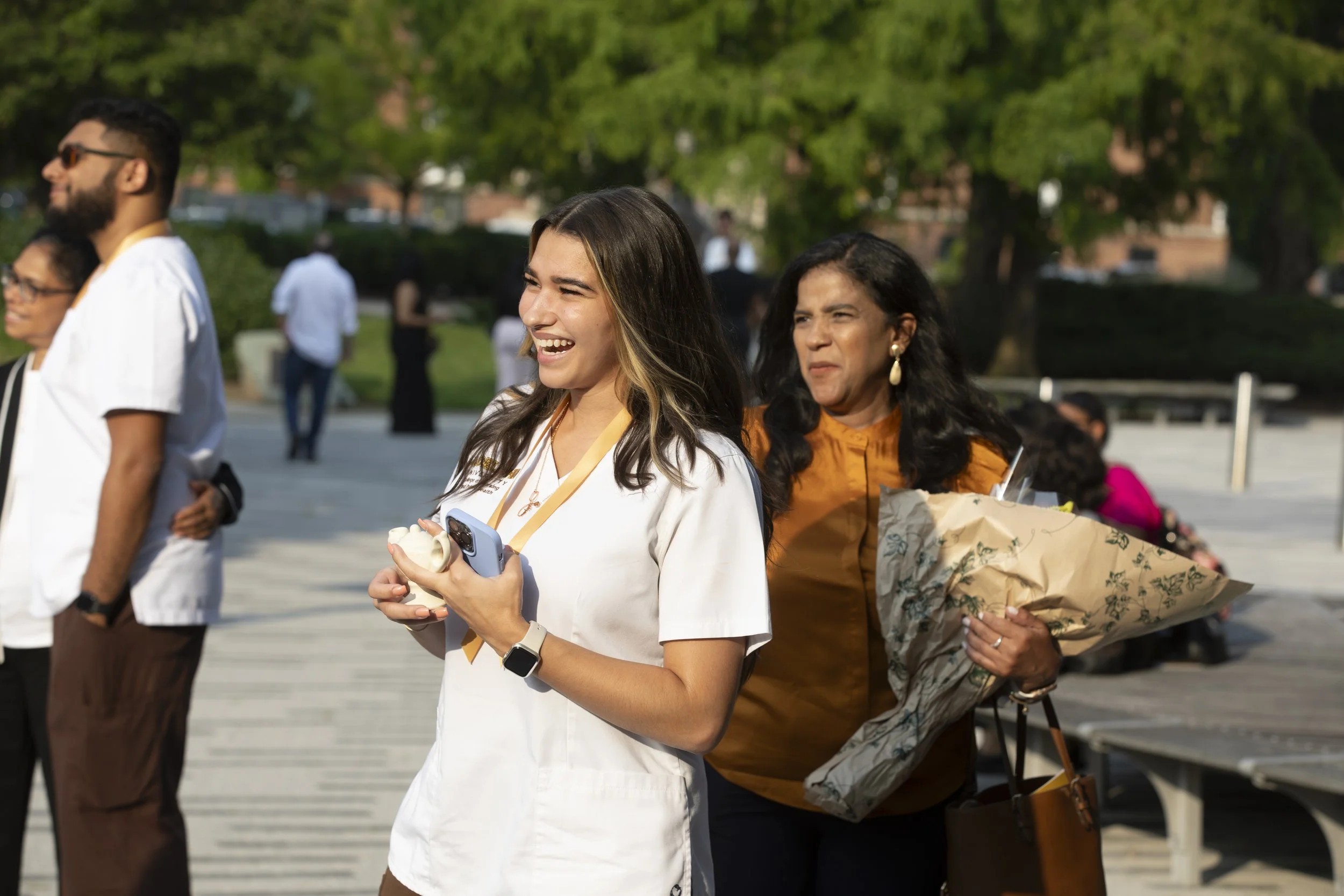 People attending an outdoor event, including a smiling young woman in a white shirt holding a phone and an older woman in an orange top carrying a large bouquet of flowers.