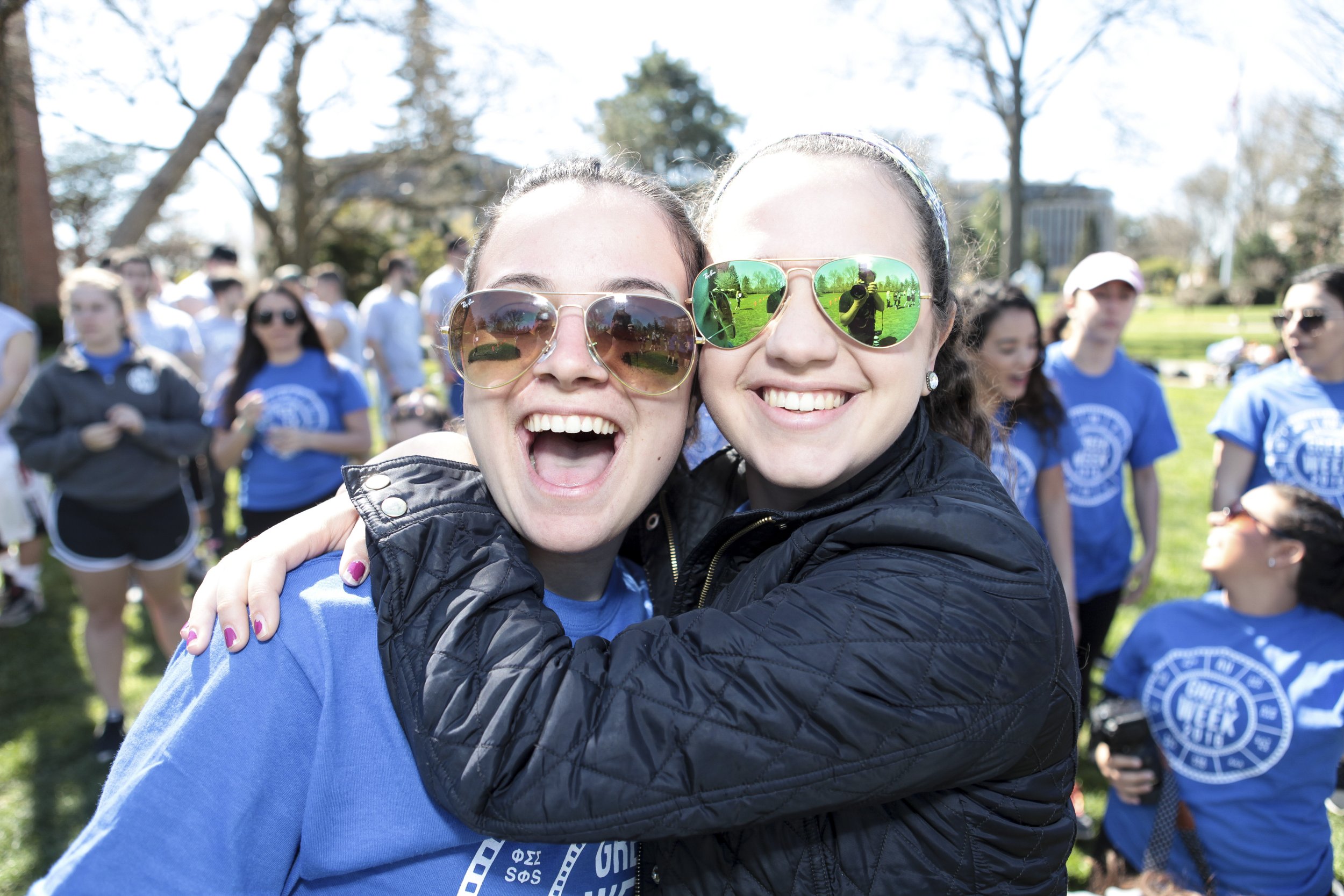 Two women smiling and hugging at an outdoor event on a sunny day, with a crowd of people in the background, some wearing blue event T-shirts and sunglasses.