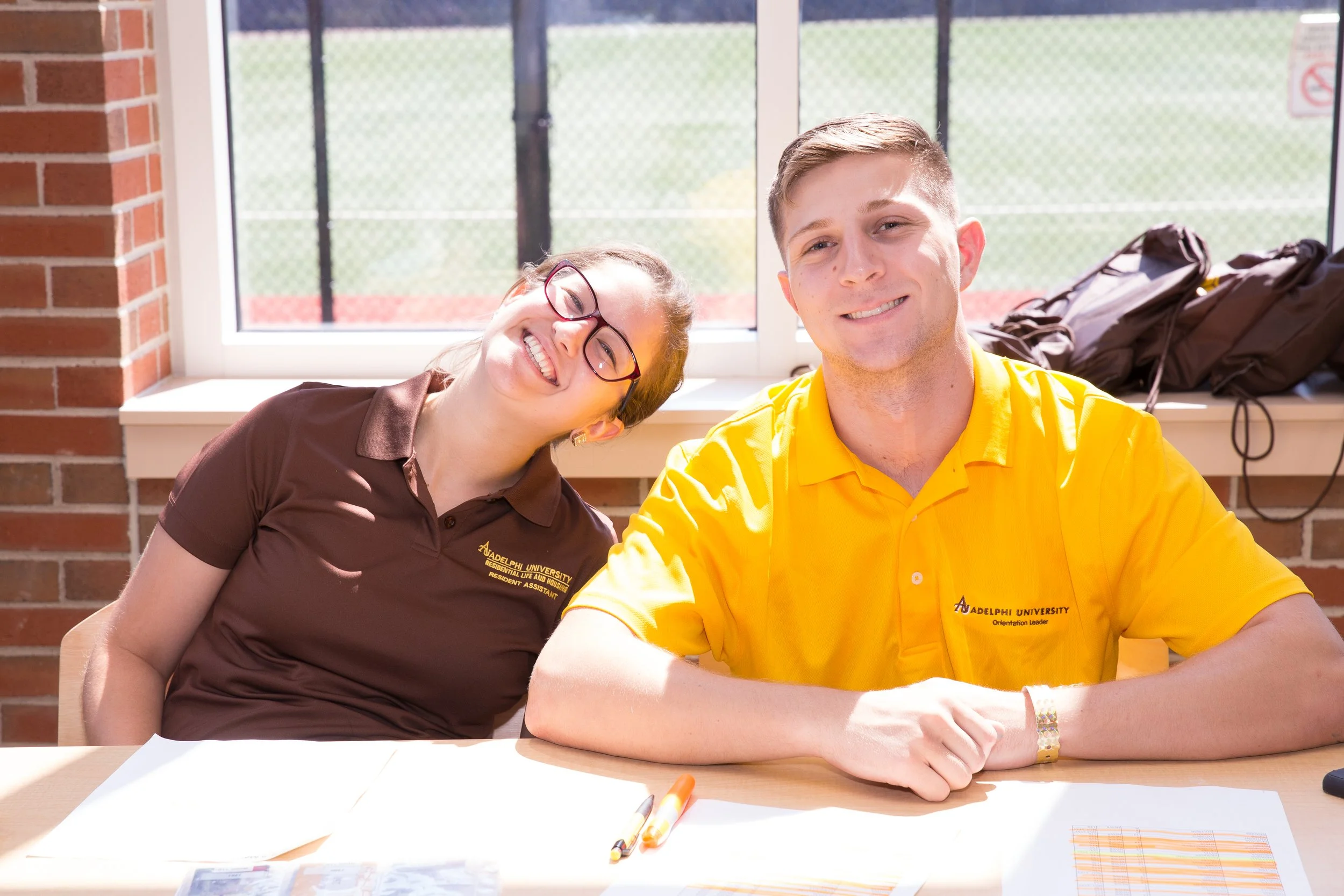 Two smiling students sitting at a table, one wearing a brown polo shirt and the other wearing a yellow polo shirt, with backpacks behind them and papers on the table.