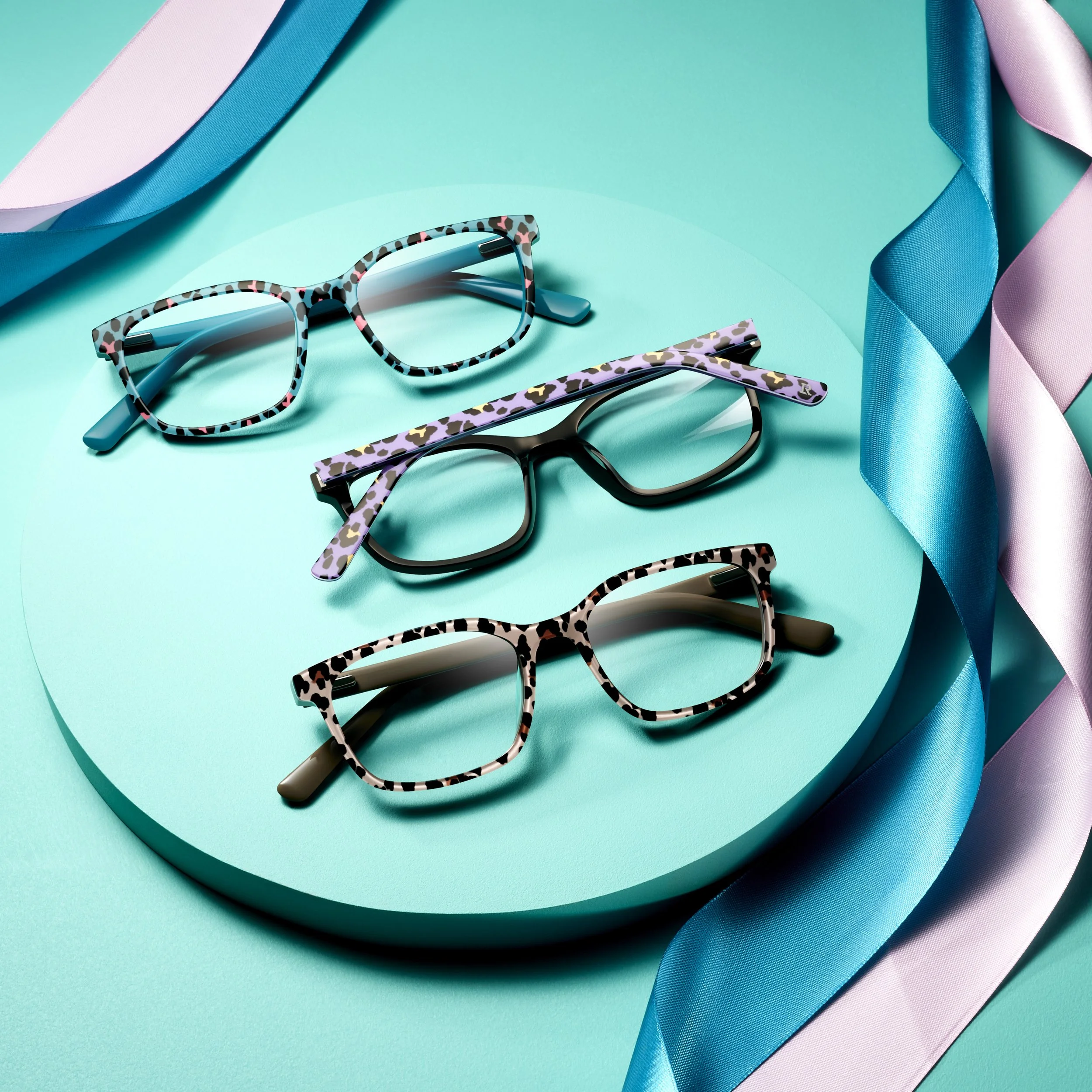 Three pairs of eyeglasses with colorful, patterned frames arranged on a turquoise circular platform, with pink, blue, and white satin ribbons framing the scene.