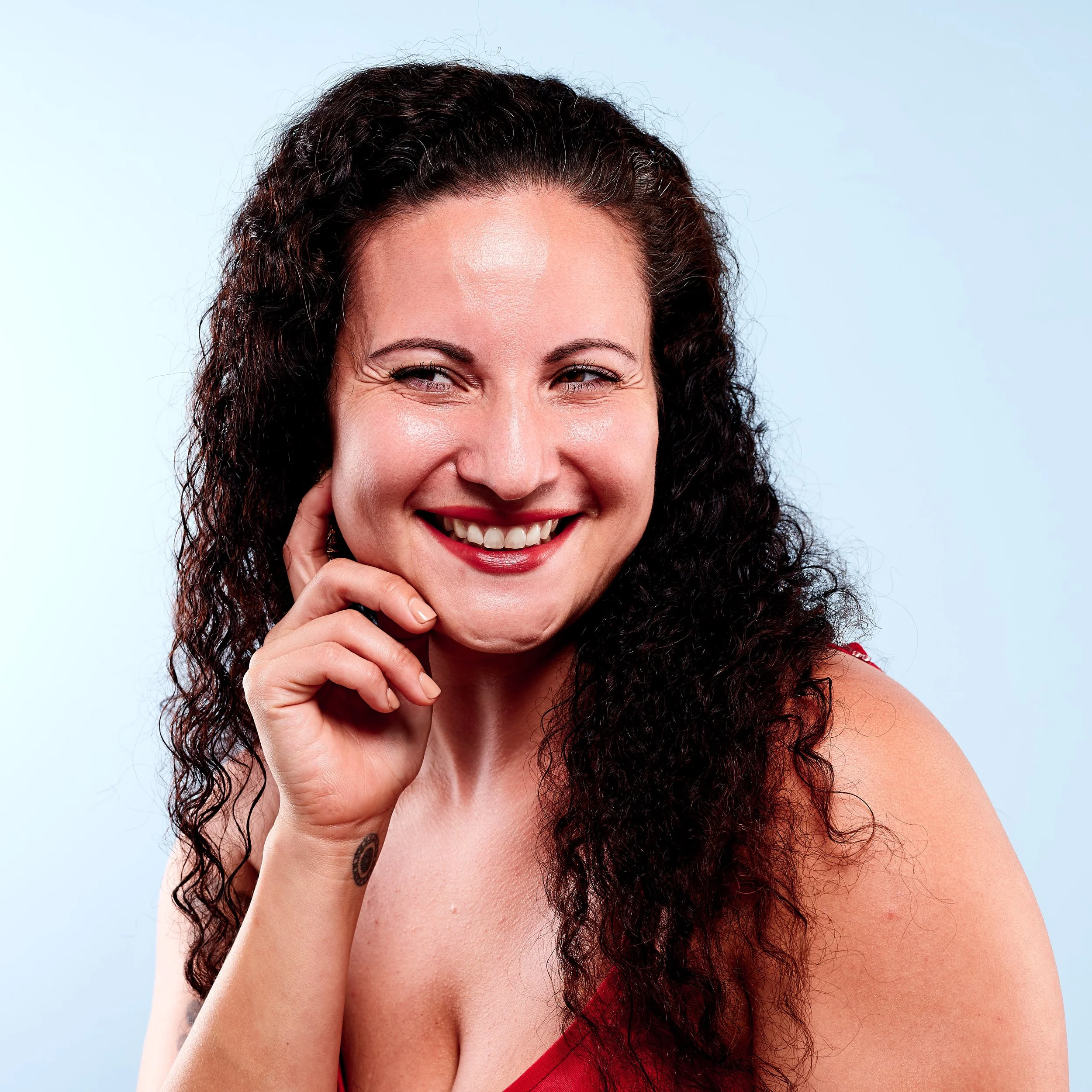 A smiling woman with curly dark hair, her eyes partially closed, touching her cheek with her fingers, wearing a red top against a light blue background.