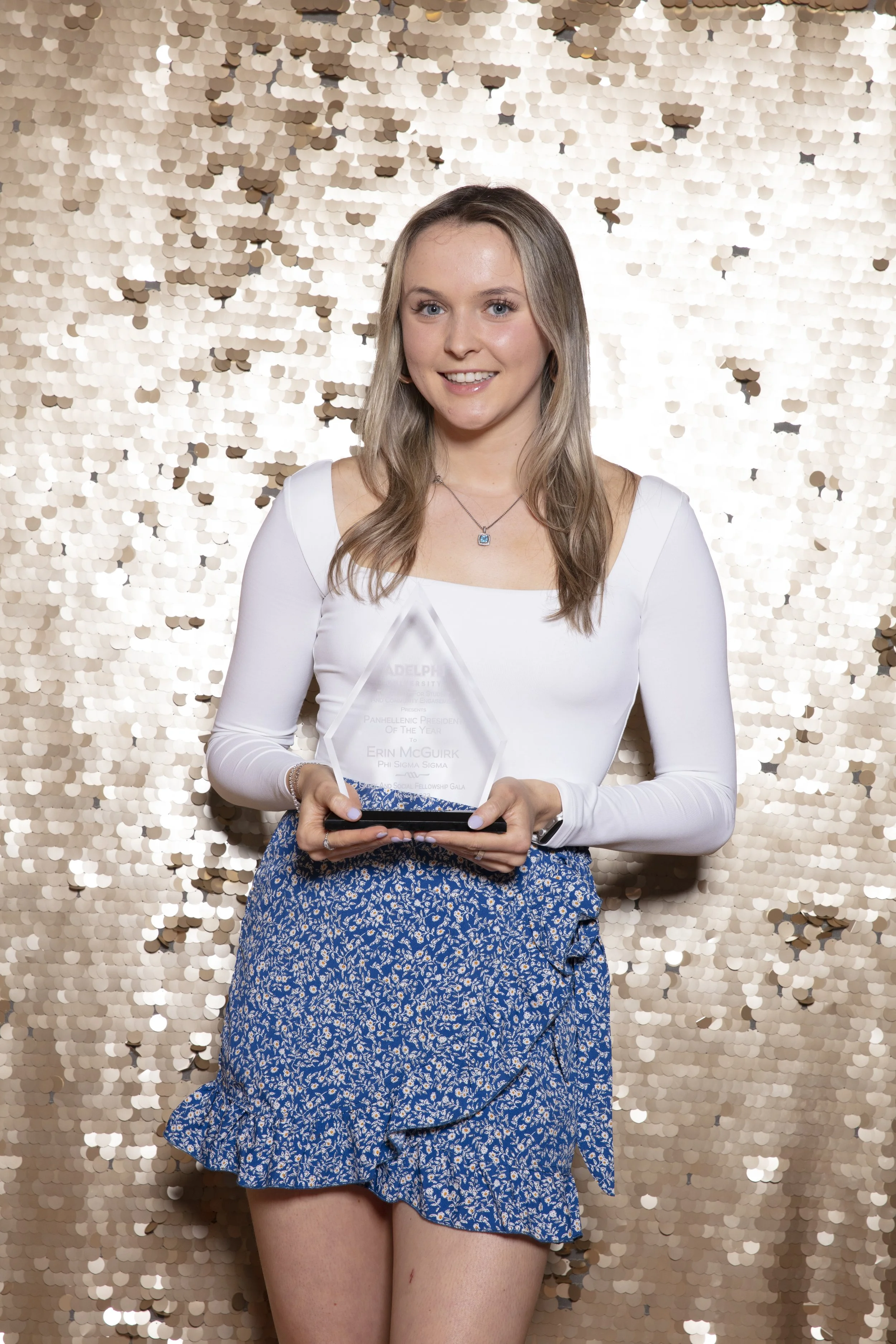 Young woman in white long-sleeve top and blue patterned skirt holding an award, standing in front of a gold sequin backdrop.