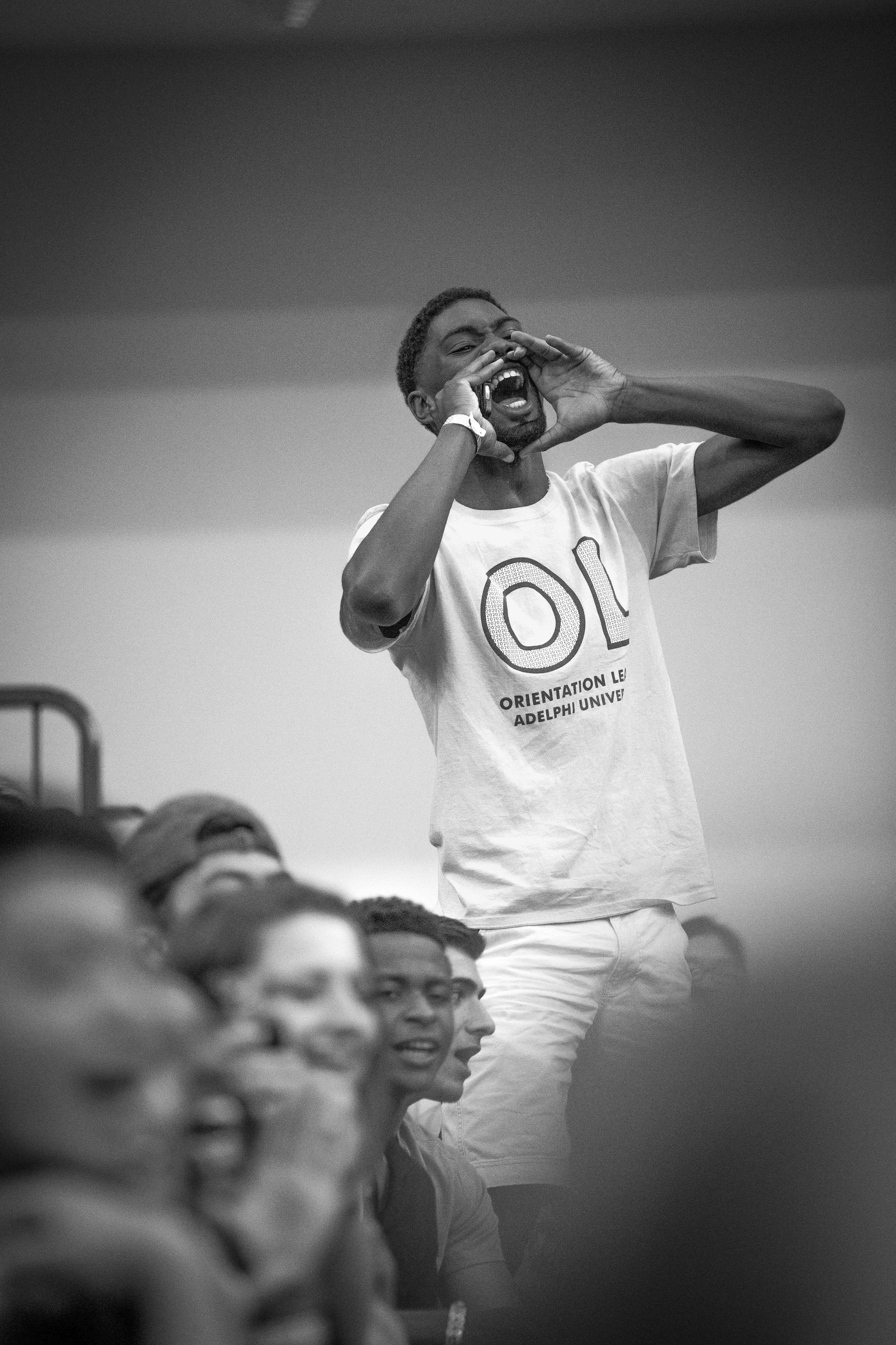 A young man shouting or calling out, standing among a seated group of people, wearing a T-shirt with 'OL' and 'Orientation Leader Adelphi University' written on it, indoors with a plain background.