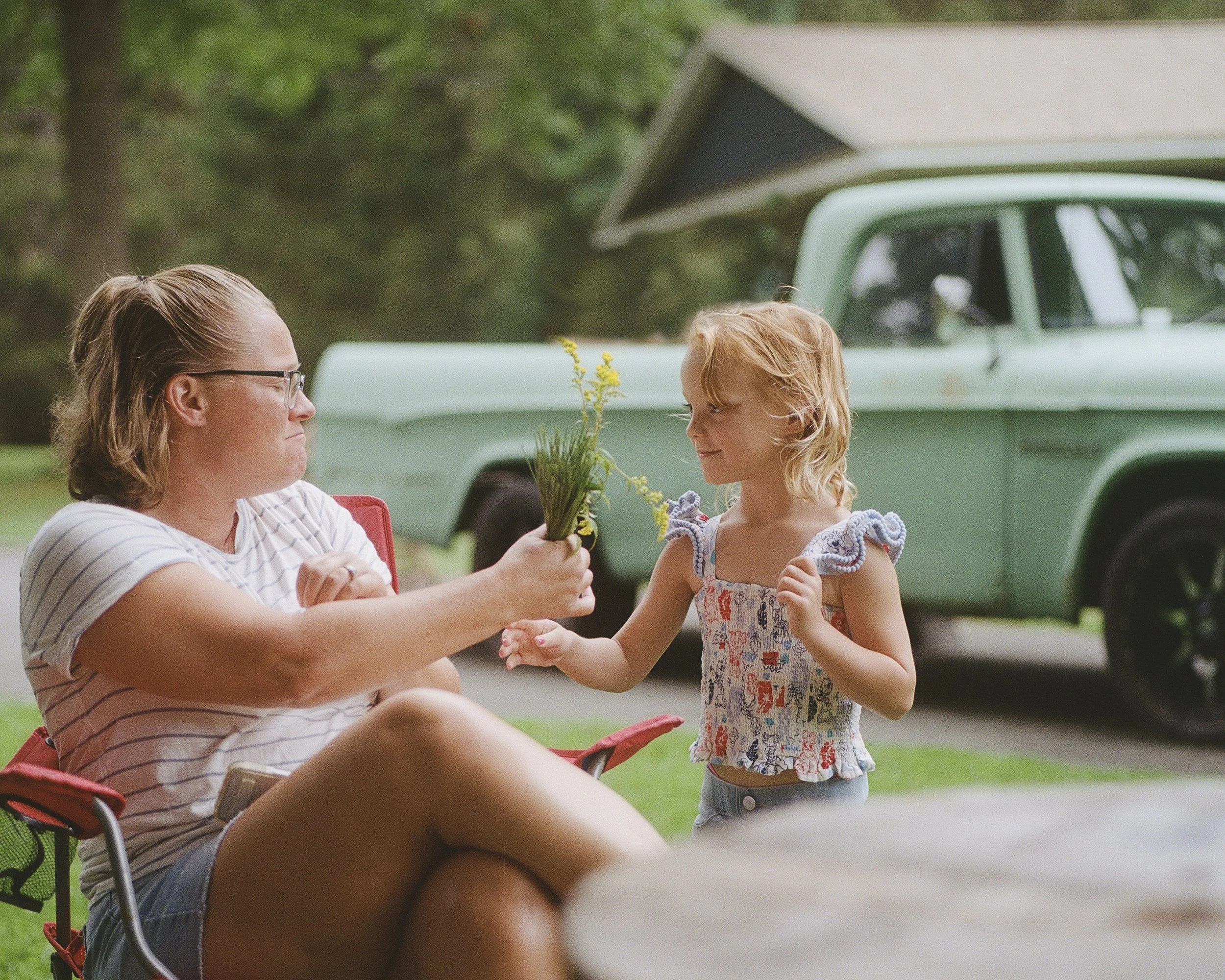 A woman with glasses and a young girl with blonde hair exchanging a small bouquet of yellow flowers outdoors near a vintage green truck.