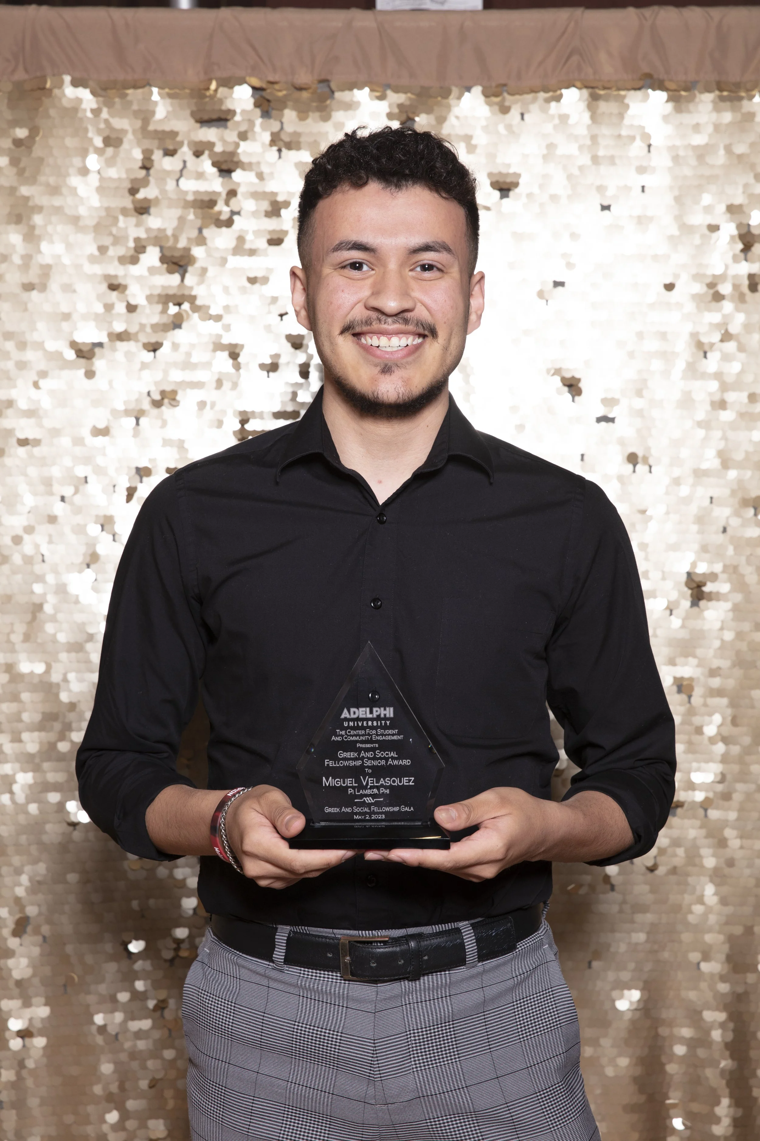 A young man in a black shirt and gray plaid pants holding a crystal award, smiling. The background features a shiny, gold sequin curtain.