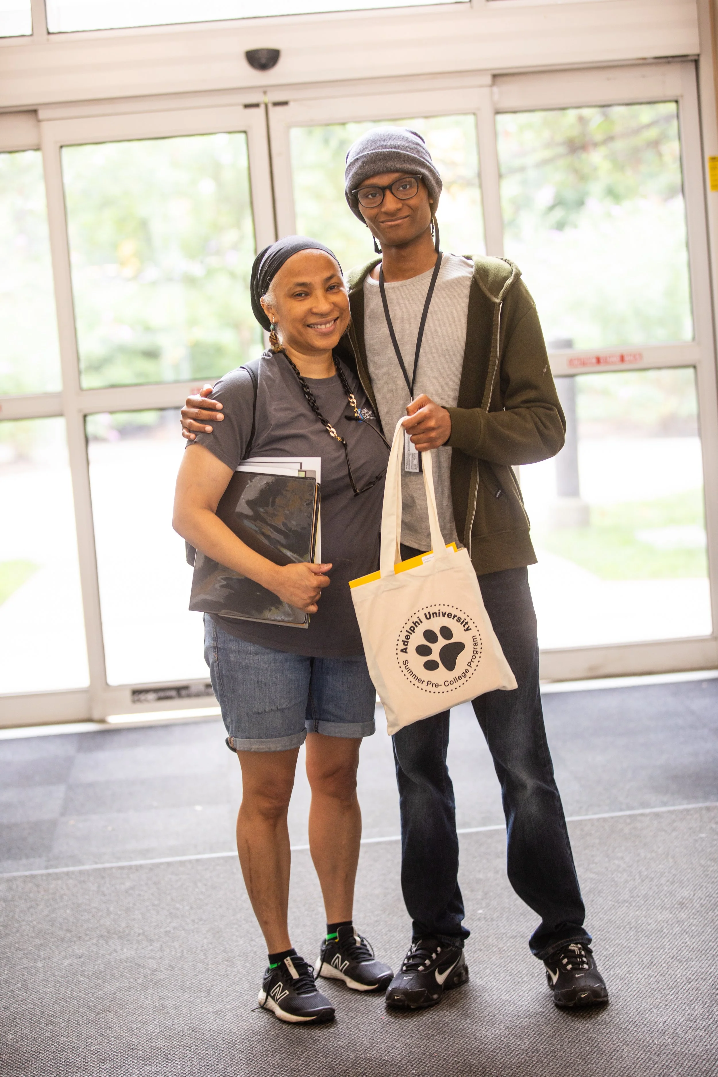 Two people standing inside near glass doors, smiling at the camera. One holds a notebook, the other holds a tote bag with 'Adelphi University Summer Pre-College Program' written on it.