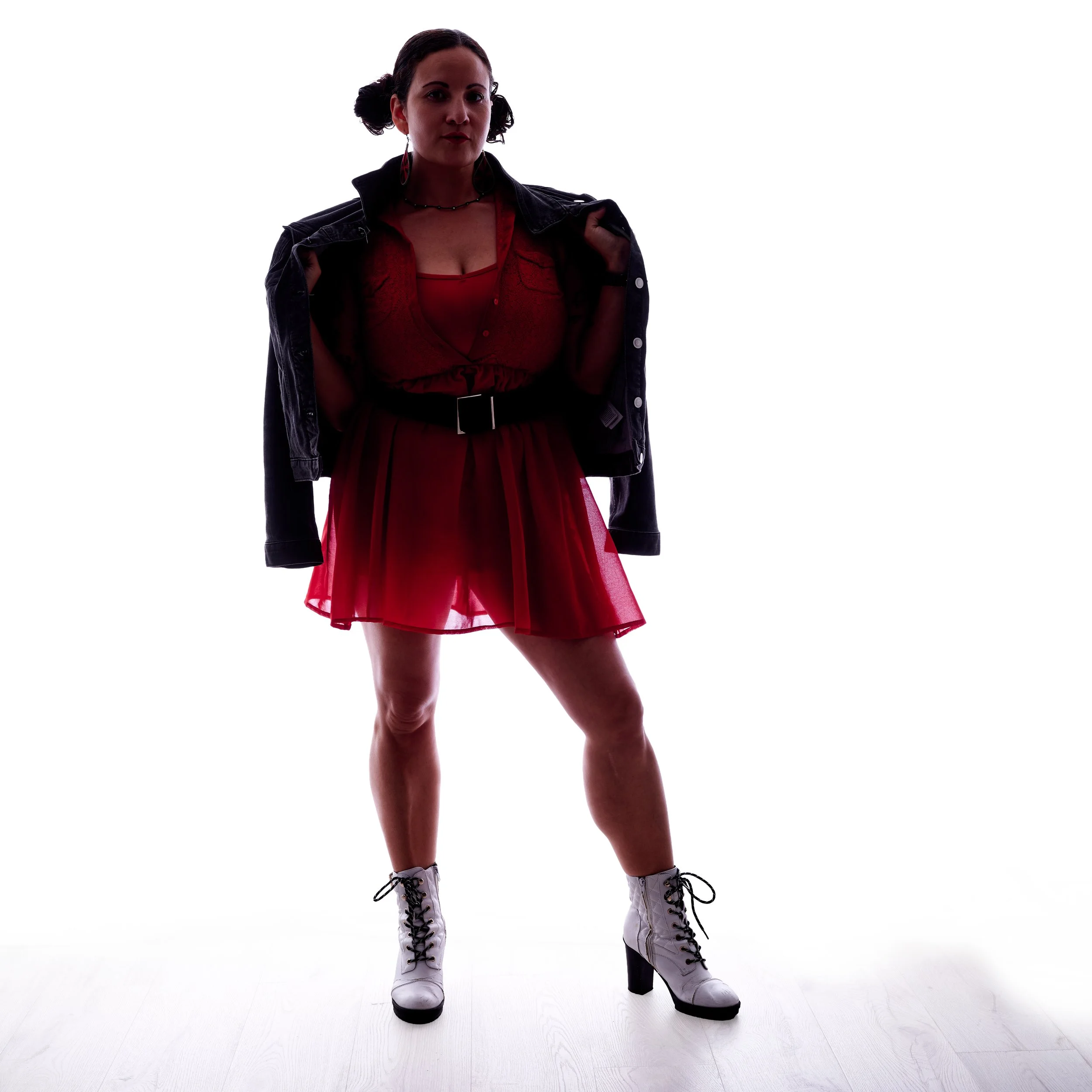 A woman standing against a white background wearing a black leather jacket, red top with lace details, a red sheer skirt, white lace-up high-heeled boots, earrings, and a necklace.