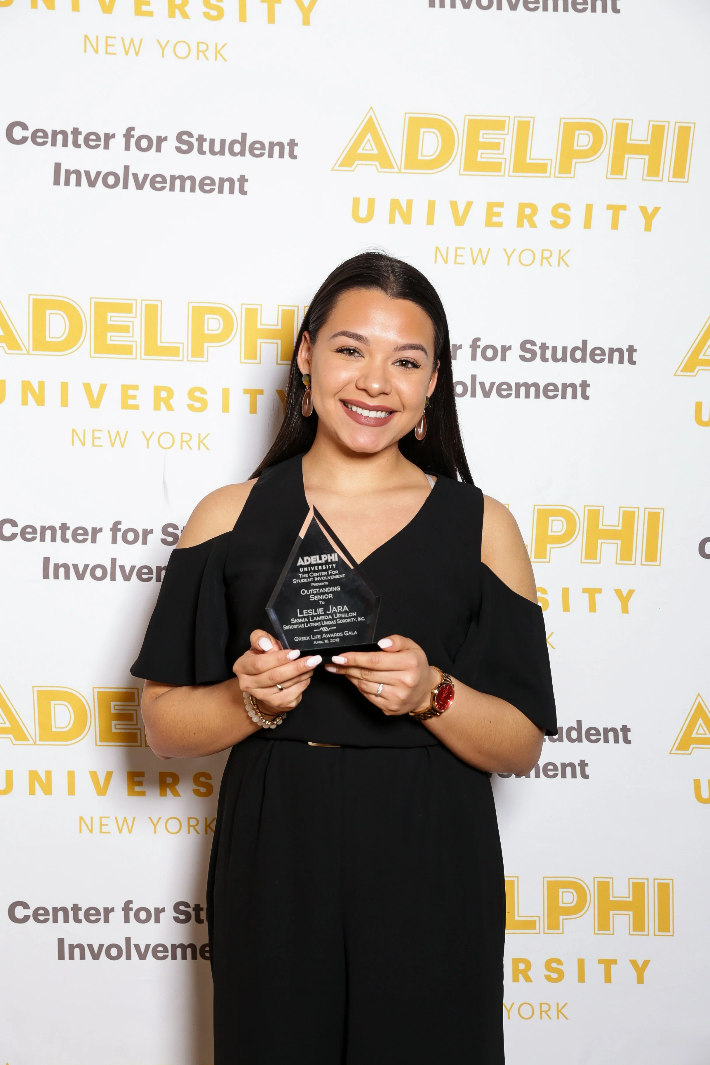 A young woman with long dark hair, wearing a black dress, holding a clear award, standing in front of a step-and-repeat banner with yellow and gray text for Adelphi University and Center for Student Involvement.
