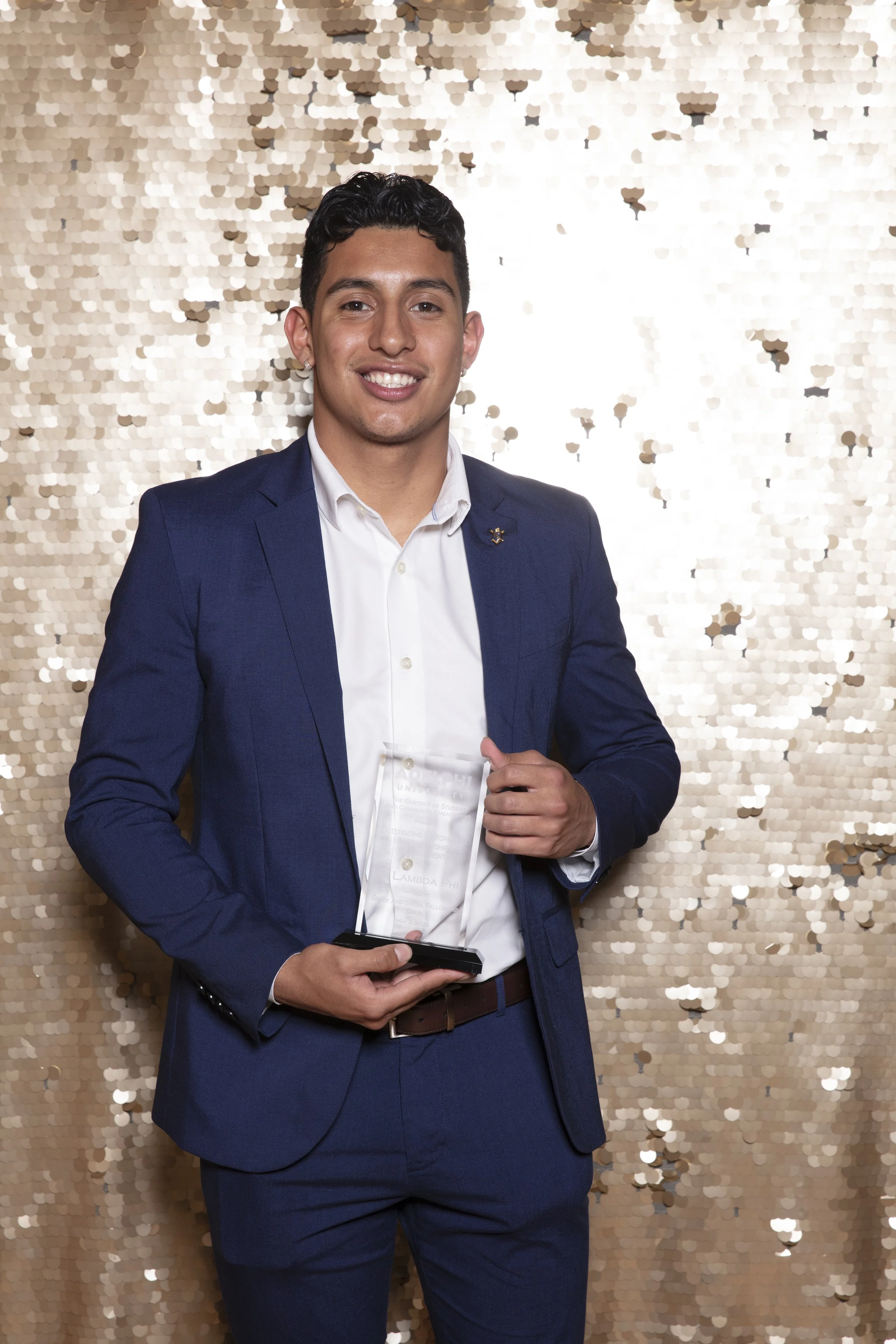 Young man in blue suit holding an award, standing in front of a gold sequin backdrop at a formal event.