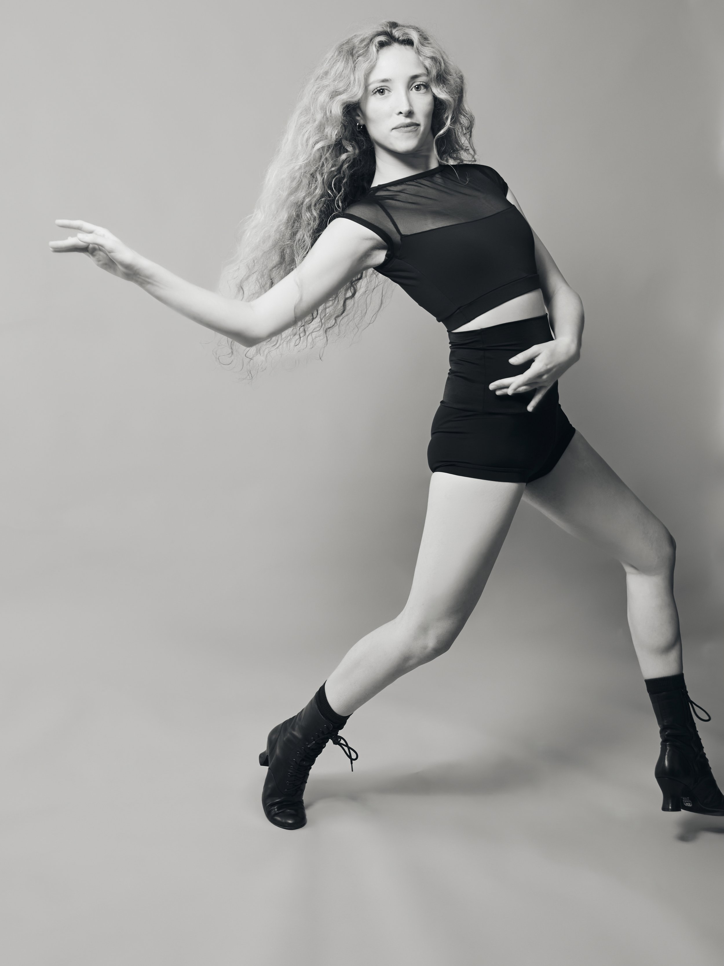A woman with curly hair dressed in a black sheer top and high-waisted shorts performing a dance pose against a plain background.