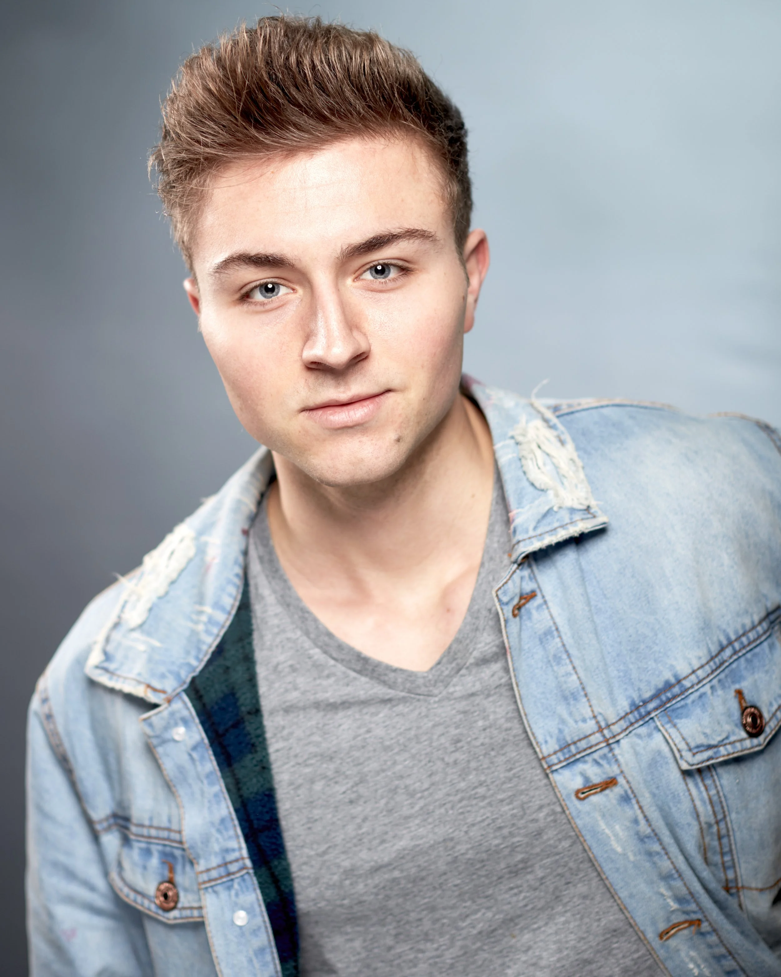 A young man with short brown hair and blue eyes wearing a gray t-shirt and a distressed denim jacket, standing against a gray background.