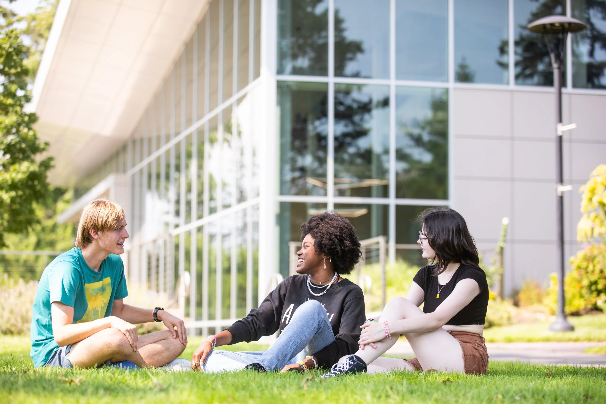Three young people sitting on grass outside and talking, with a modern glass building in the background.
