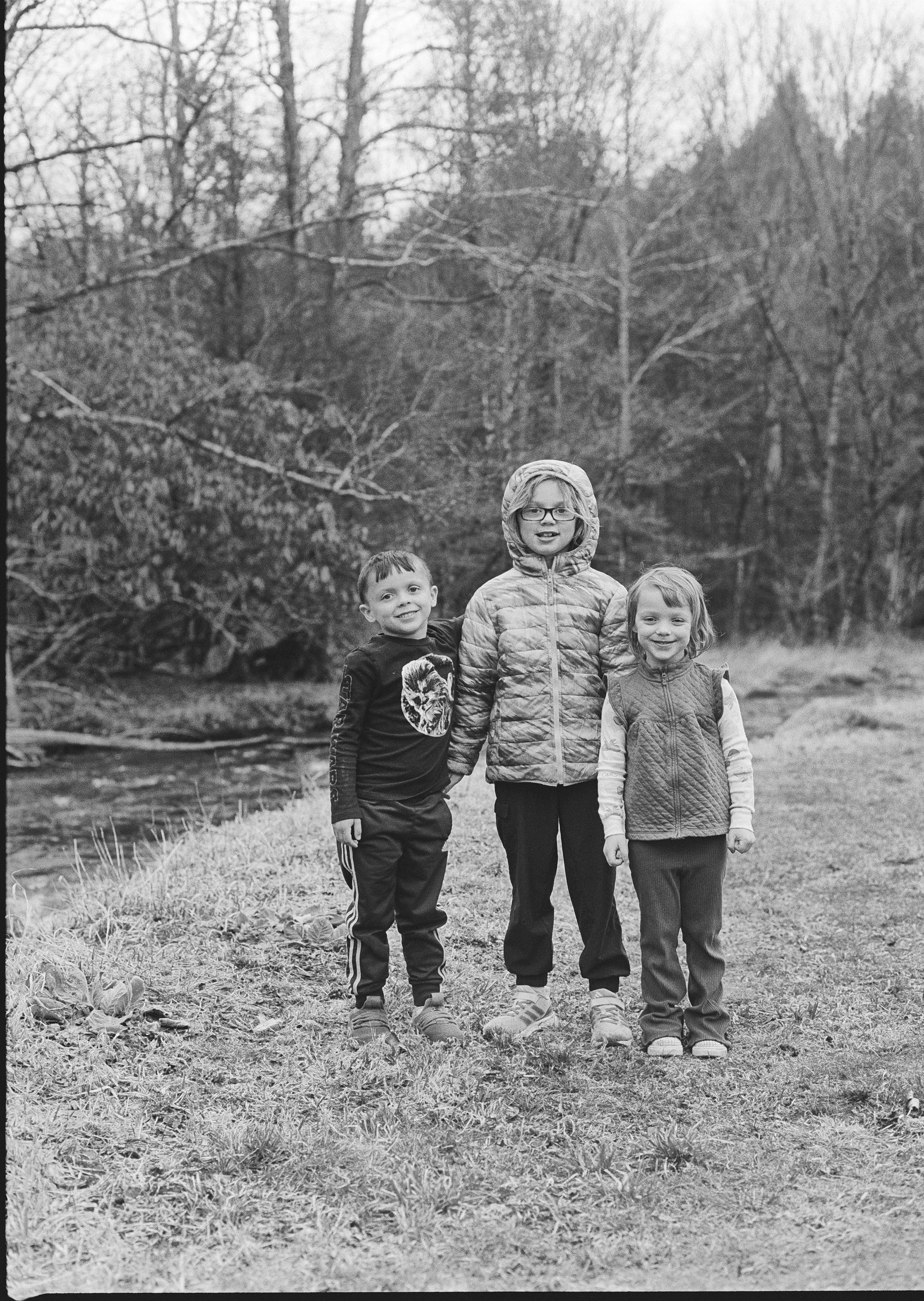 Three children standing outdoors near a creek, with trees in the background, in a black and white photograph.