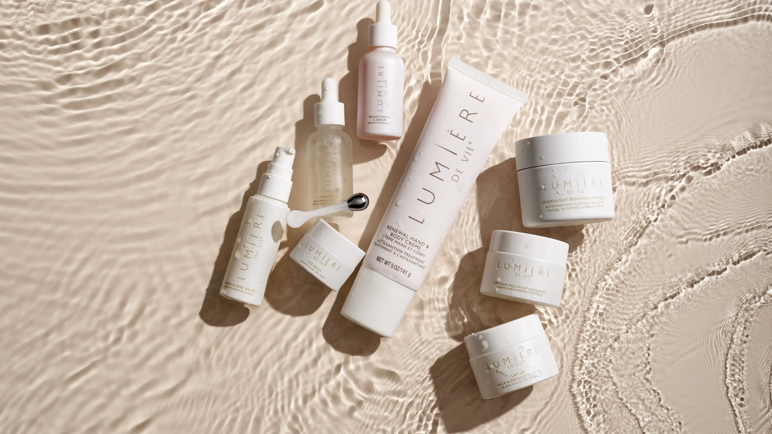 Collection of skincare and beauty products from Lumière de Vie arranged on a sandy surface, with a large tube, jars, and small bottles visible.
