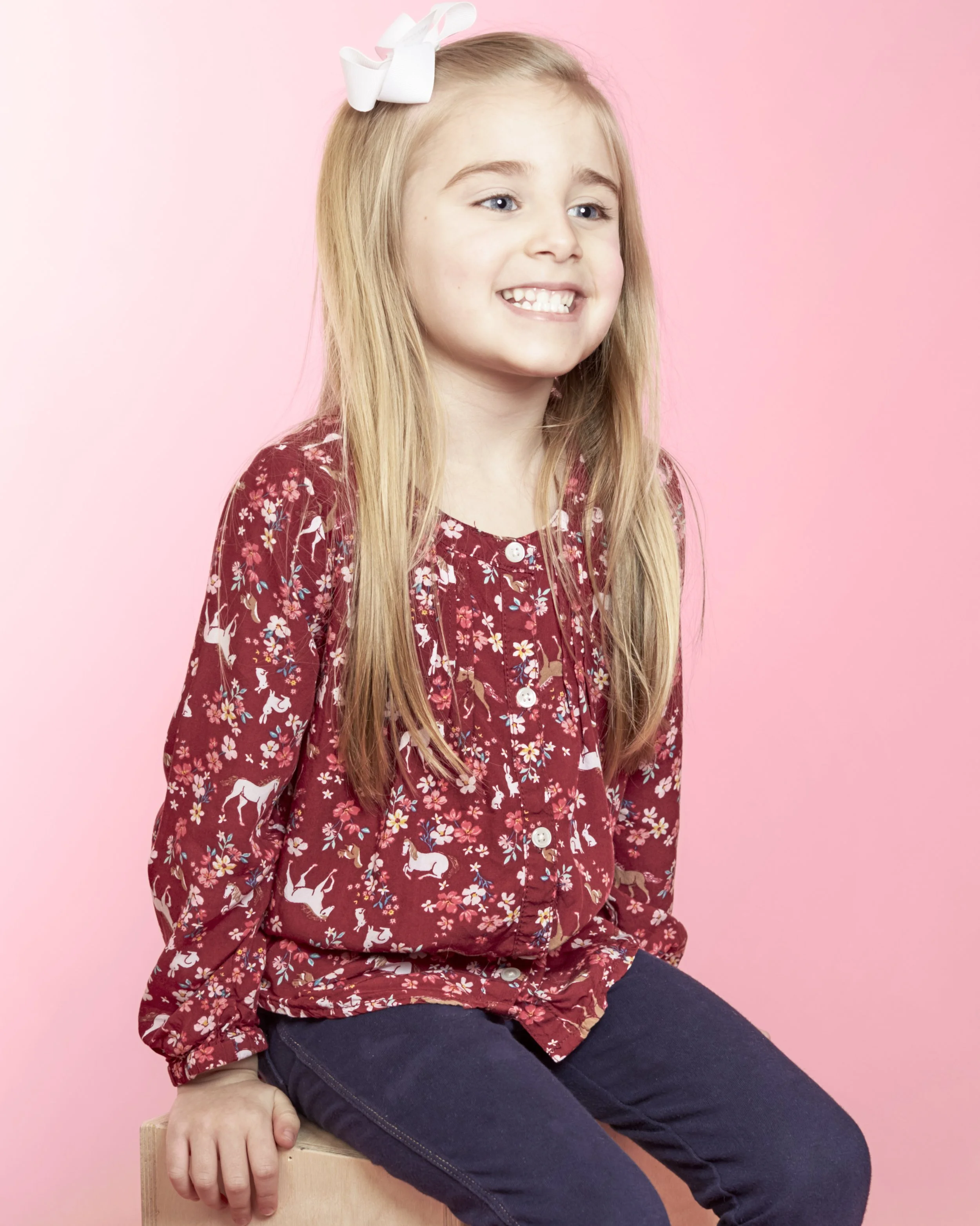 Young girl with long blonde hair, wearing a red long-sleeve shirt with a white floral and animal pattern, black pants, and a white bow in her hair, smiling while sitting on a wooden block against a pink background.