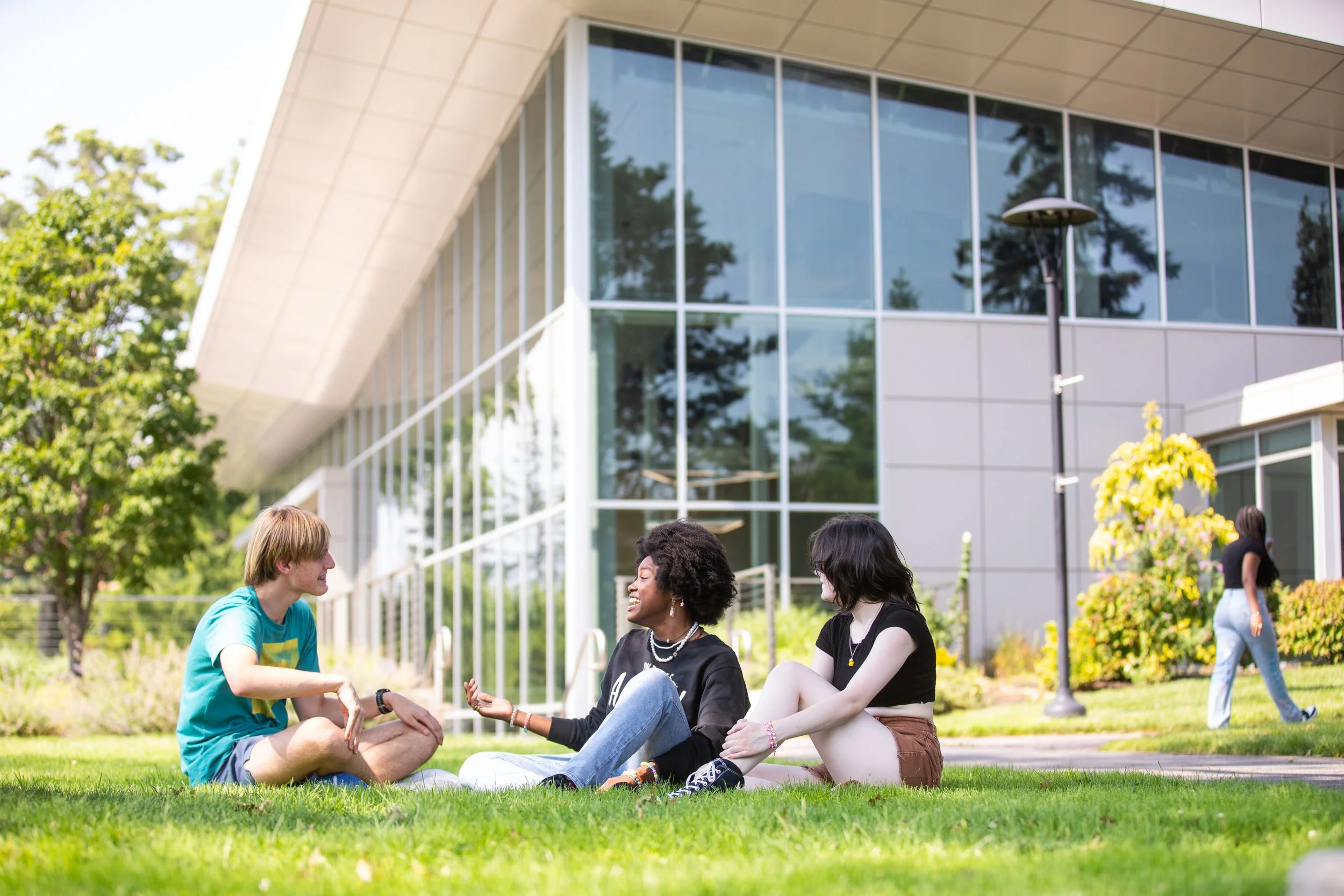 Three young people sitting on the grass, engaged in conversation, with a modern glass building in the background and another person walking nearby.
