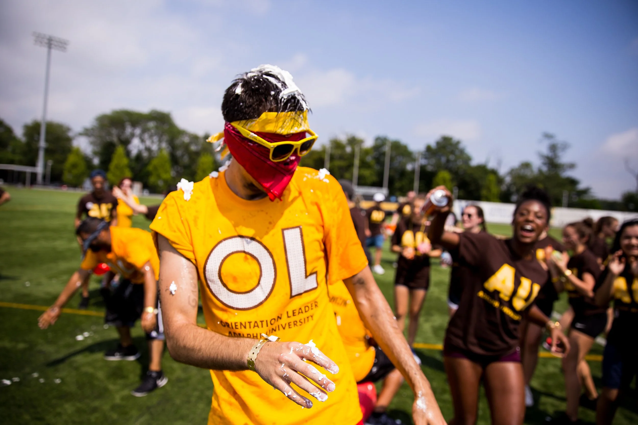 A group of people celebrating on a sports field, with one person in the foreground wearing sunglasses, a red bandana covering their face, and a yellow shirt with the letters 'O' and 'L' visible, covered in pie or cream.