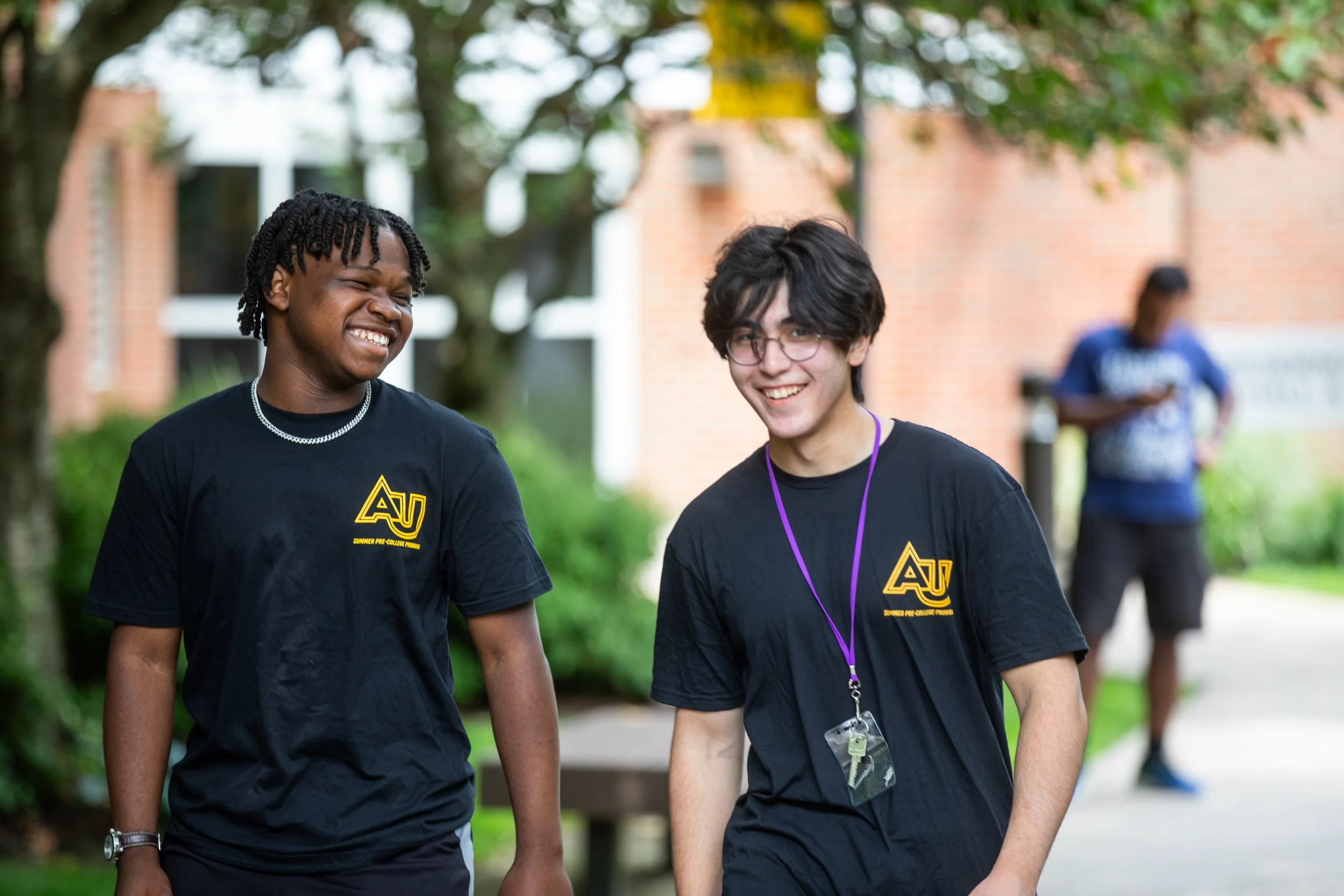 Two young men walking outdoors and smiling, wearing black T-shirts with a yellow logo, with a third person blurred in the background.