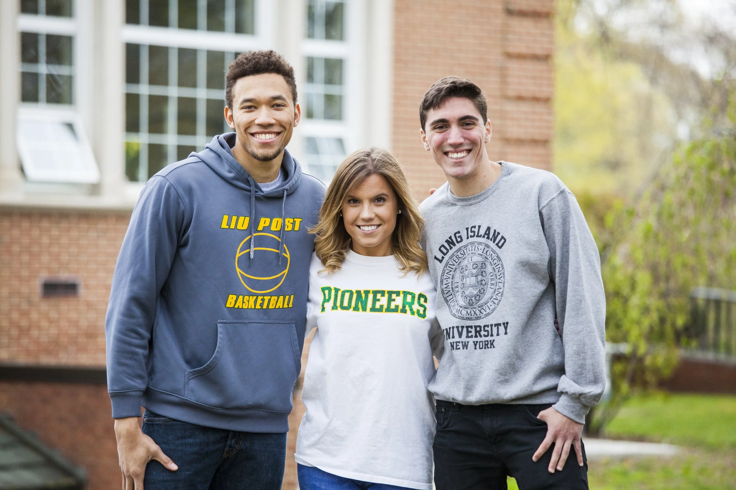 Three smiling young adults standing outdoors, with a brick building and trees in the background. The person on the left is wearing a blue hoodie with yellow text and a basketball logo. The woman in the middle is wearing a white t-shirt with green tex