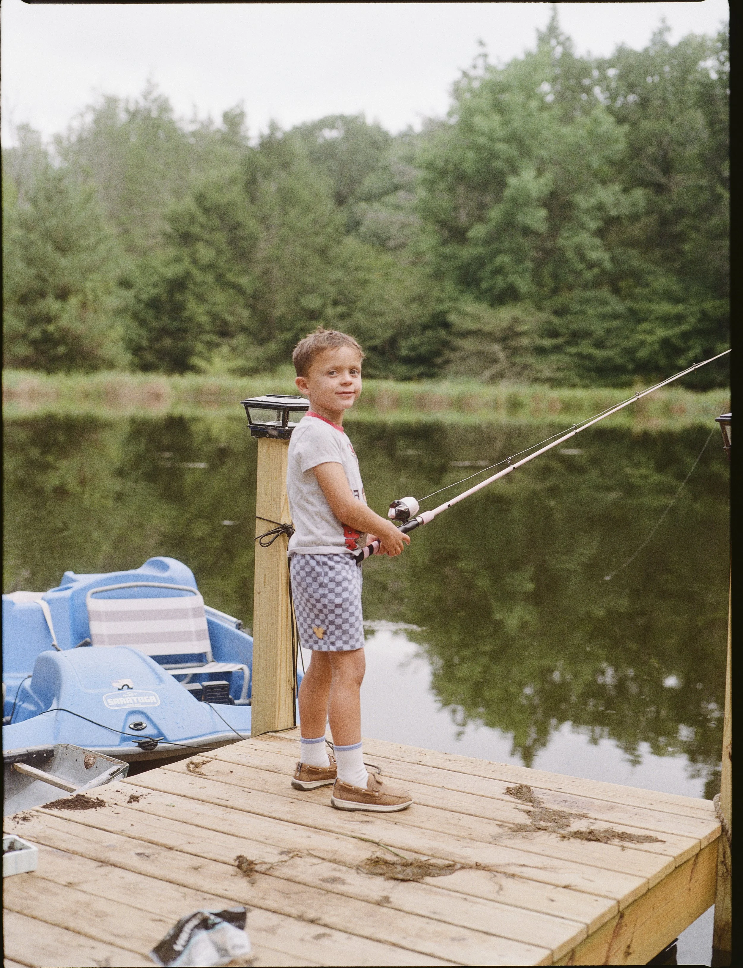 A young boy fishing off a wooden dock by a lake surrounded by trees during daytime.