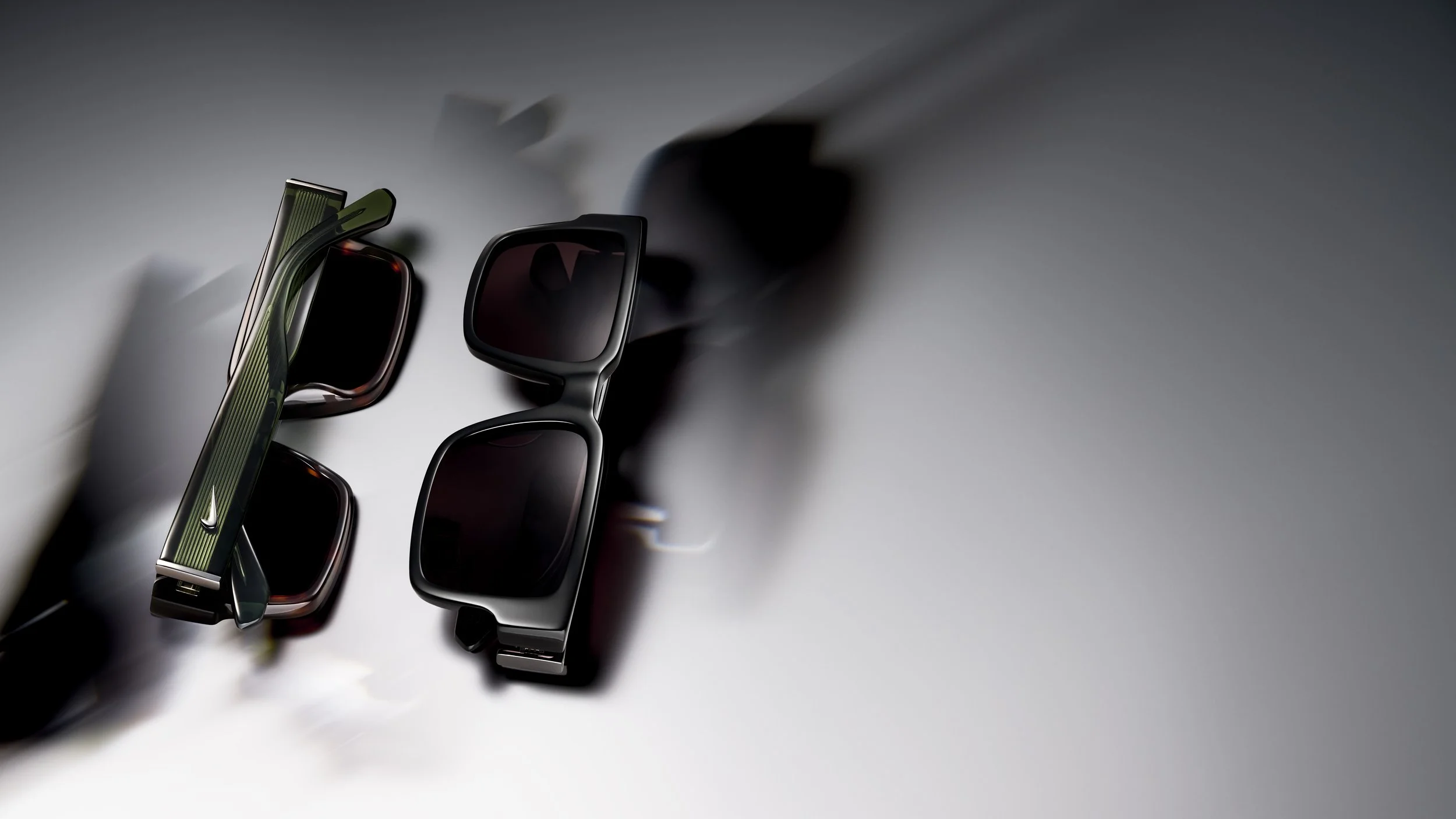 Various sunglasses and eyeglasses arranged on a white surface with shadows cast across them.