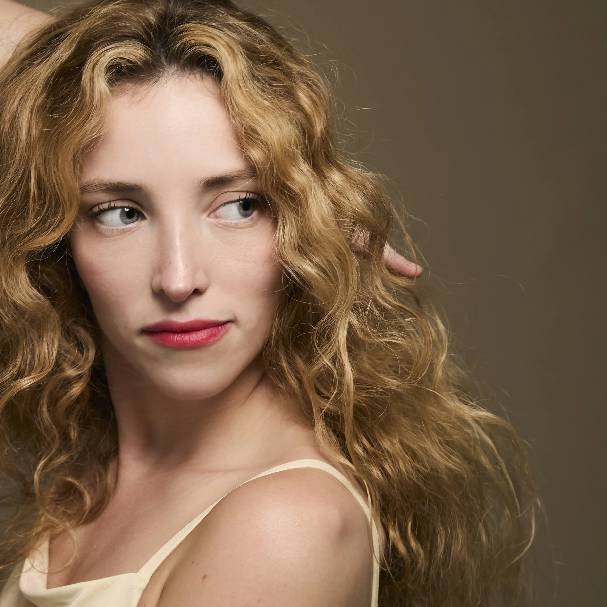 Close-up of a woman with curly red hair and fair skin, wearing a cream-colored top, showing part of her face, eye, and shoulder.