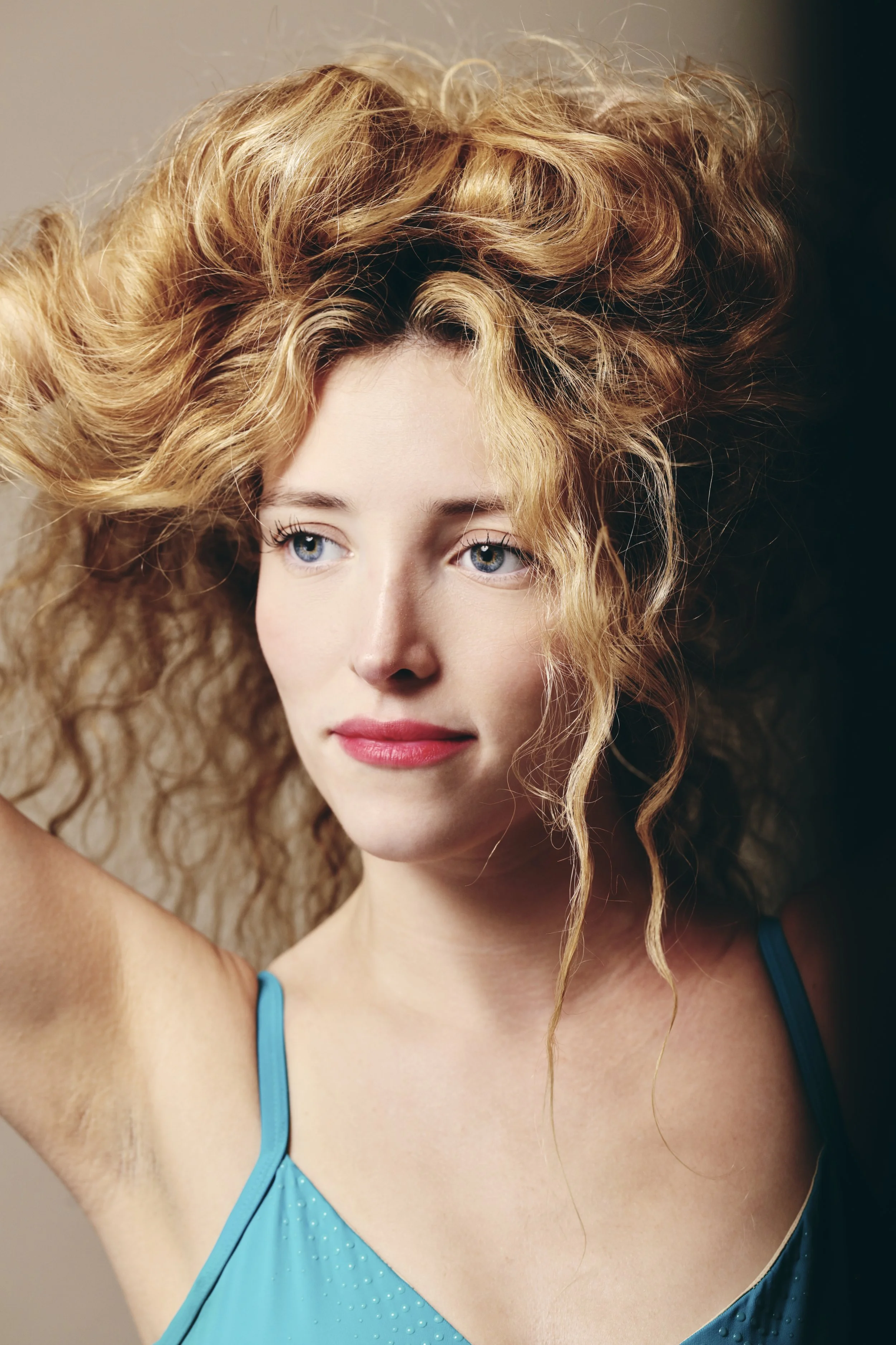 Close-up of a young woman with curly red hair, blue eyes, and pink lipstick, wearing a blue spaghetti strap top.