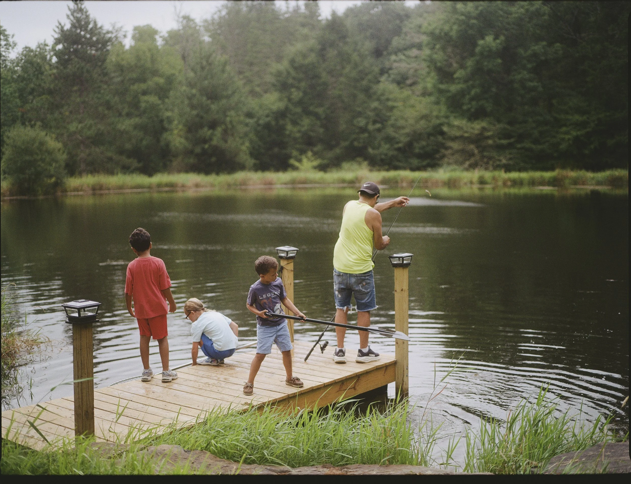 A group of people, including children and adults, are on a small wooden dock by a lake, fishing. A man in a yellow shirt and a black cap is casting his line, while children are watching and participating. The background includes a forested area with 