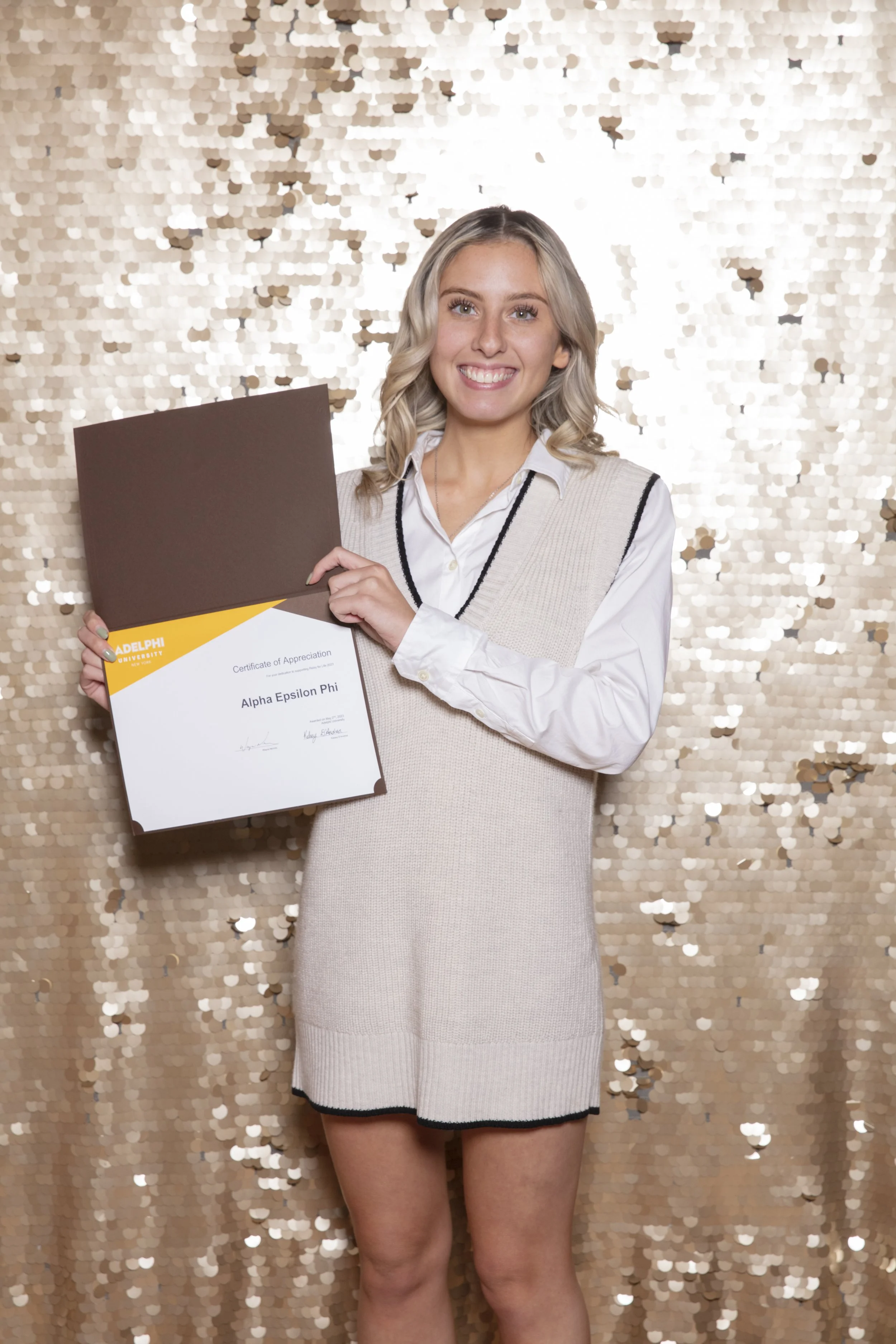 Young woman smiling and holding a certificate of appreciation from Adelphi University, standing in front of a gold sequin backdrop.
