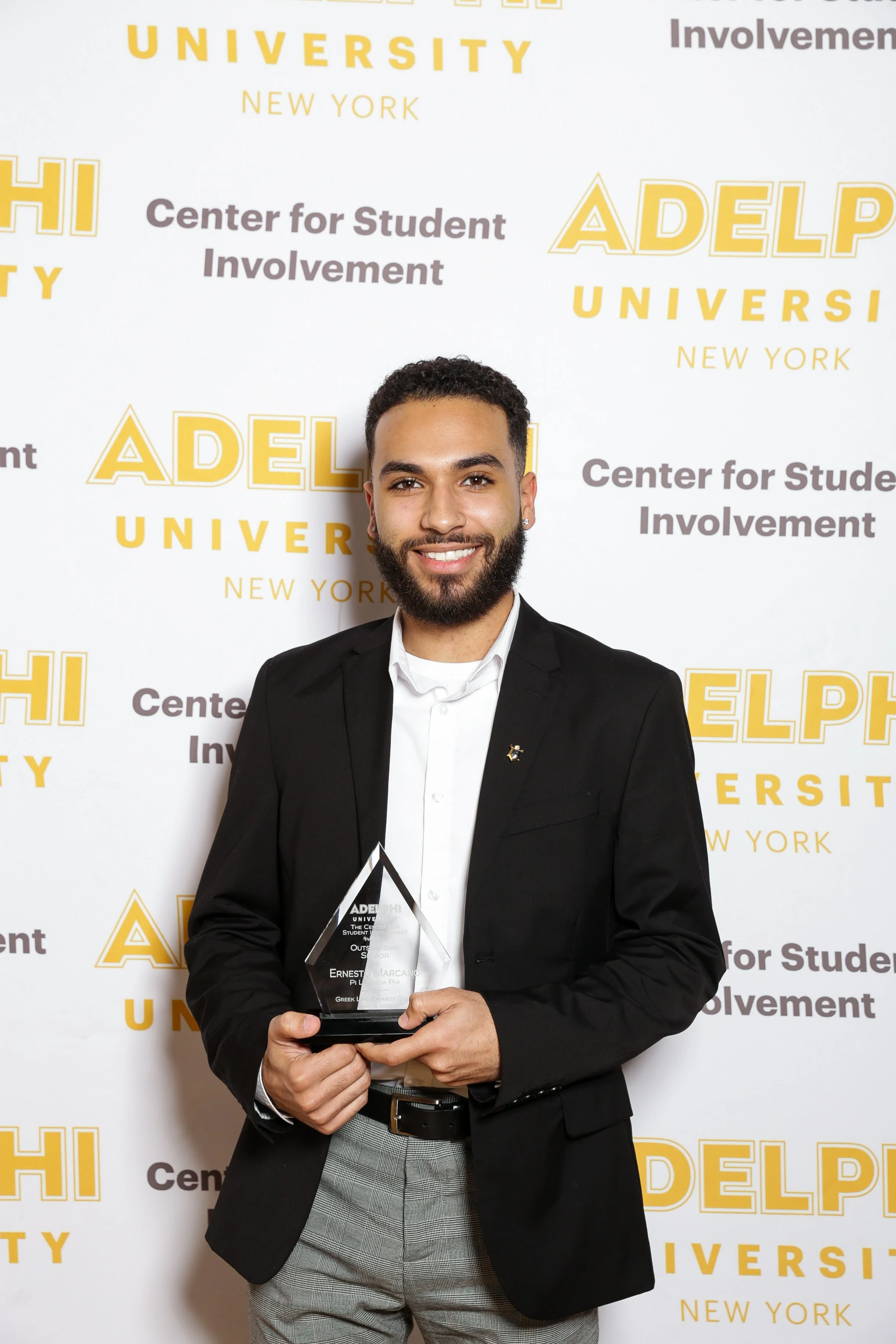 A smiling man in a black blazer and gray pants holding a glass award, standing in front of a step-and-repeat banner with logos that read 'ADELPHI UNIVERSITY' and 'Center for Student Involvement'.