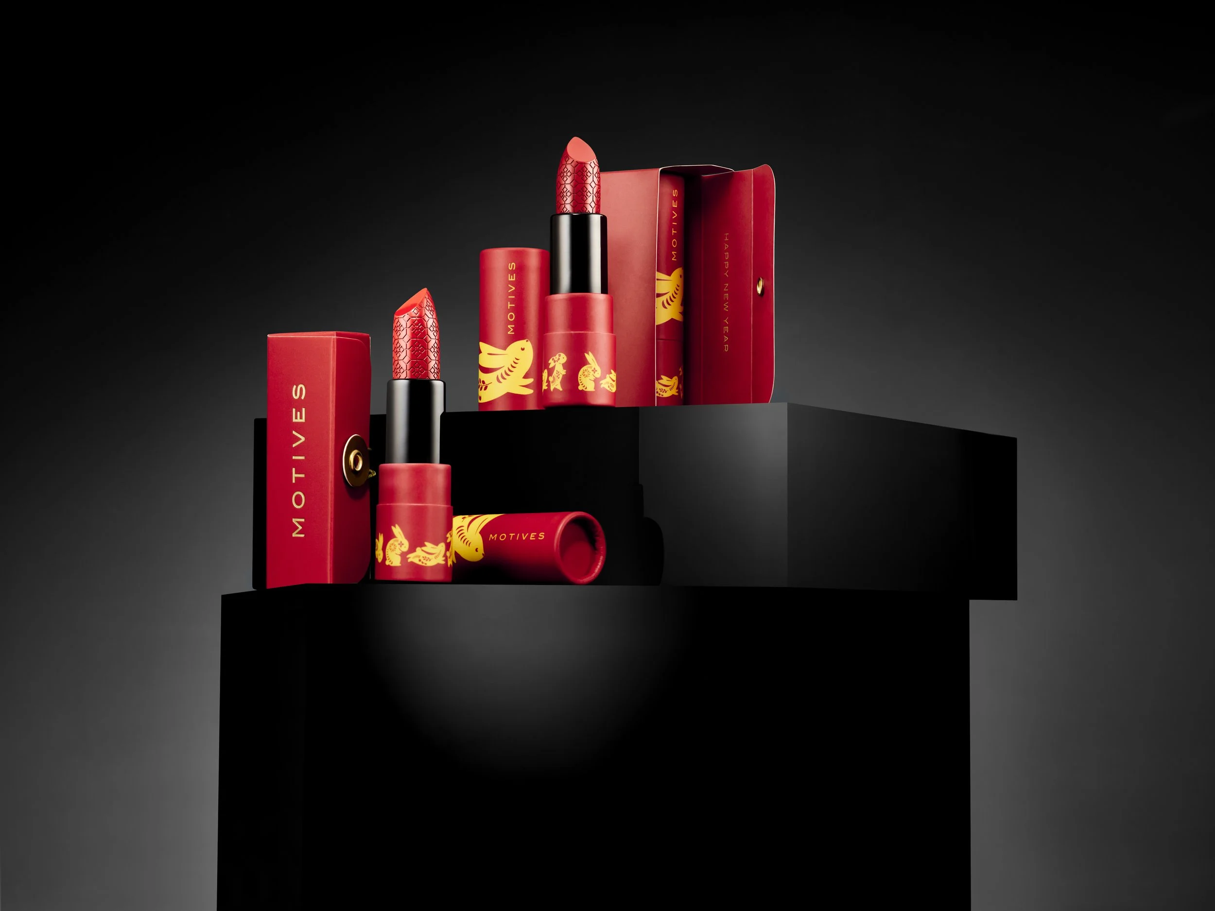 Red lipstick and lipstick cases with gold floral designs on black platforms, set against a dark background.
