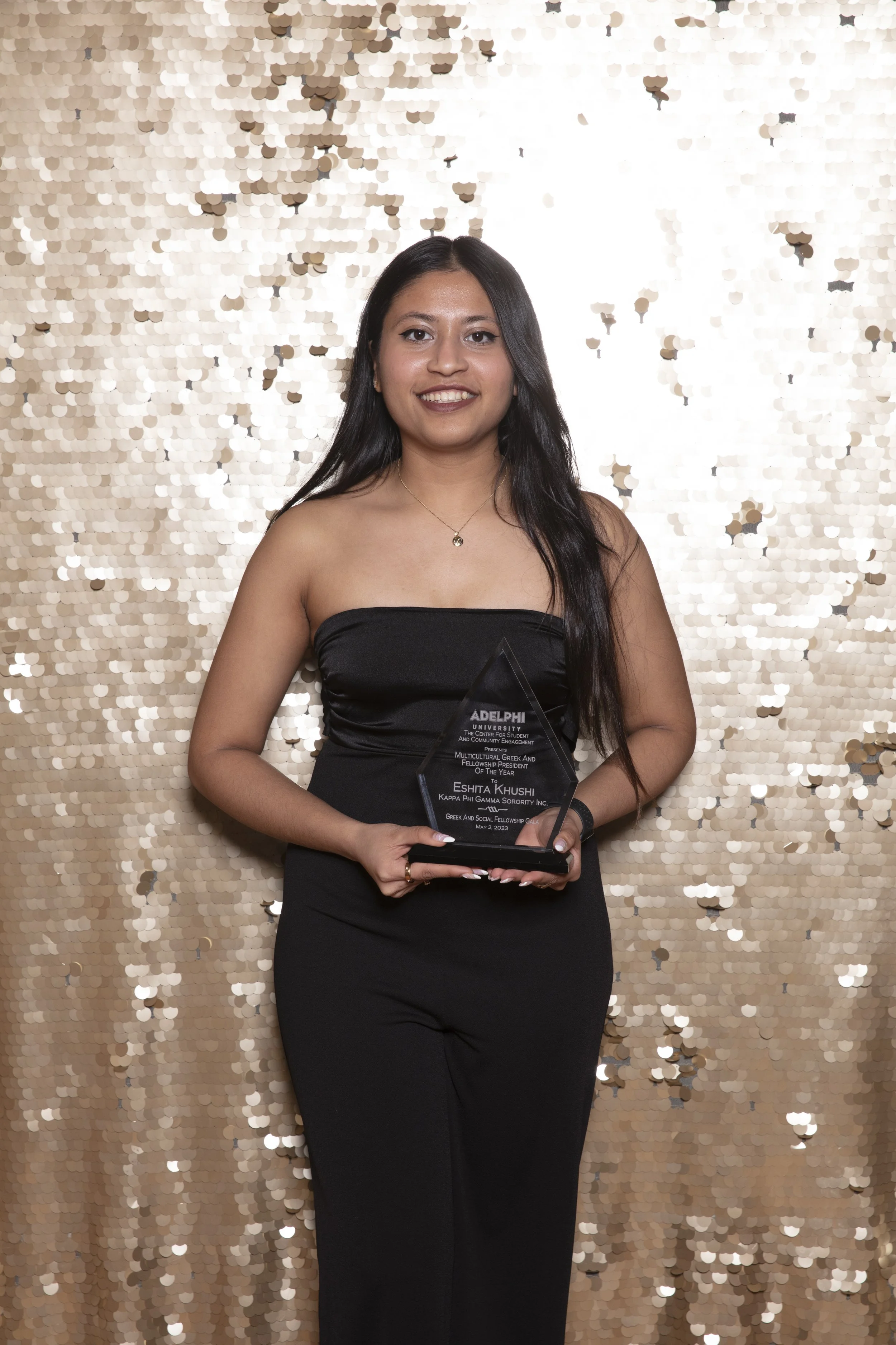 A young woman with long dark hair wearing a black strapless dress stands in front of a gold sequin backdrop, holding an award plaque for multicultural Greek and Fellowship President of the Year, dated May 2, 2023.