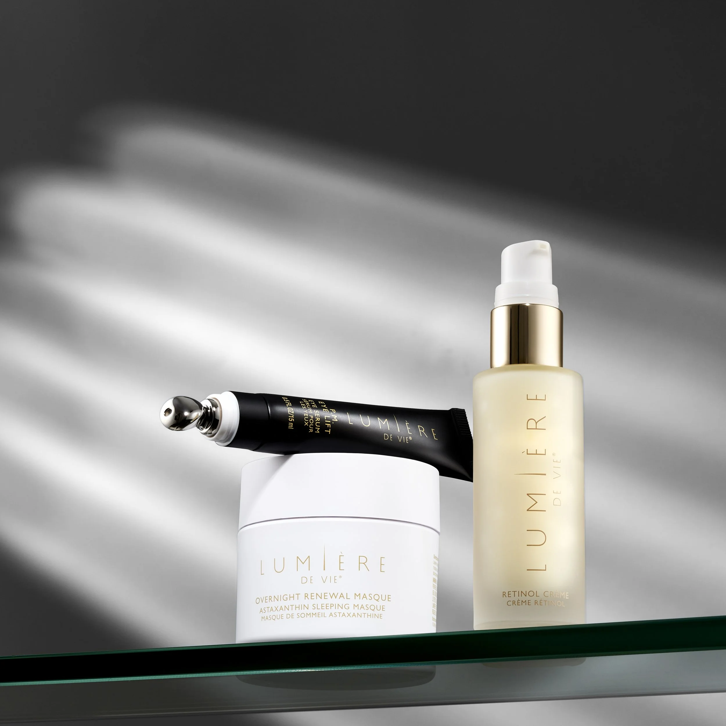 Three skincare products from Lumière de Vie are displayed, including a jar of overnight renewal masque, a retinol crème, and a black tube of eye serum with a silver applicator, on a glass shelf against a blurred background.