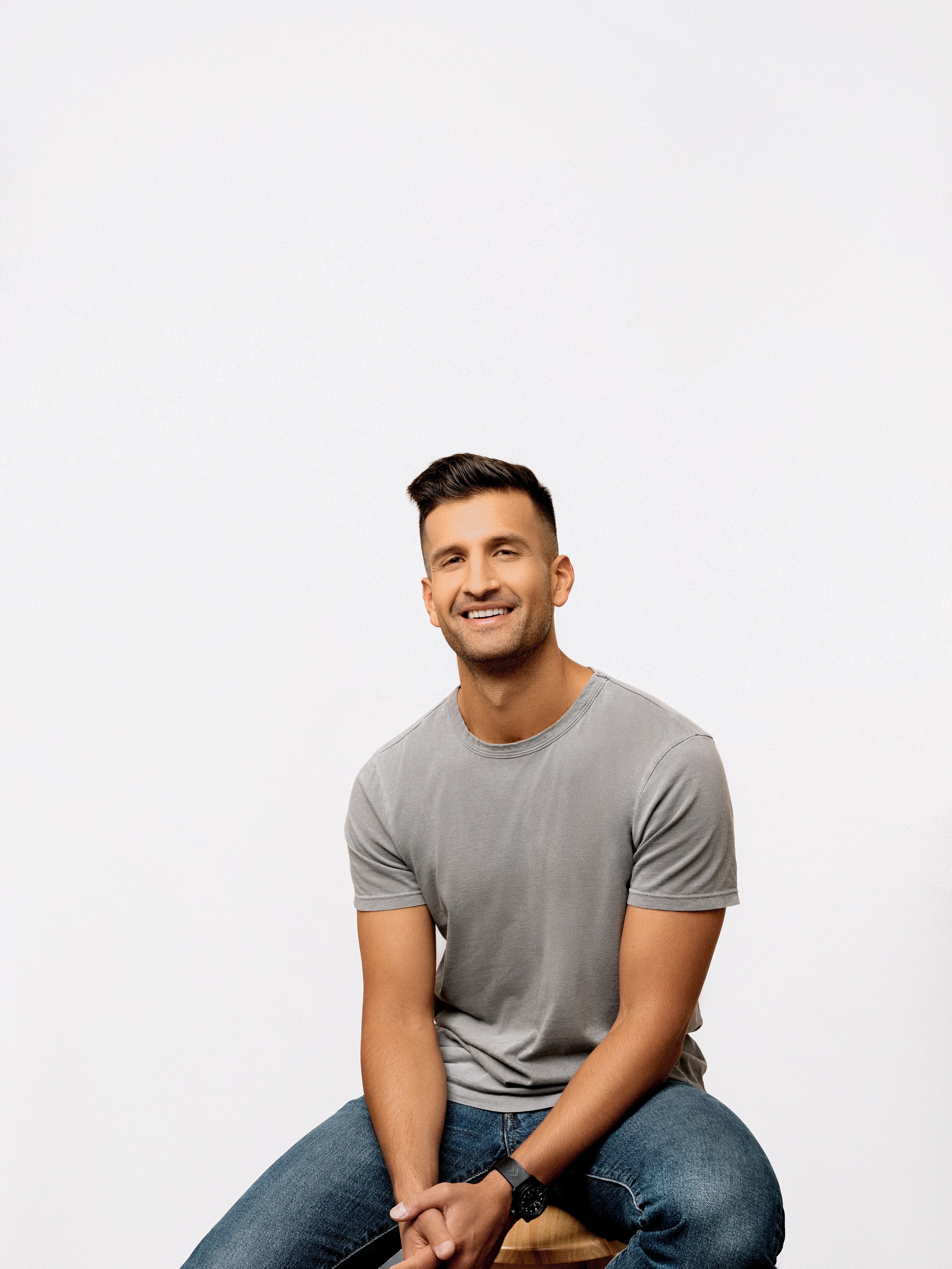 A man sitting on a stool in front of a plain white background, smiling at the camera, dressed in a gray t-shirt and jeans.