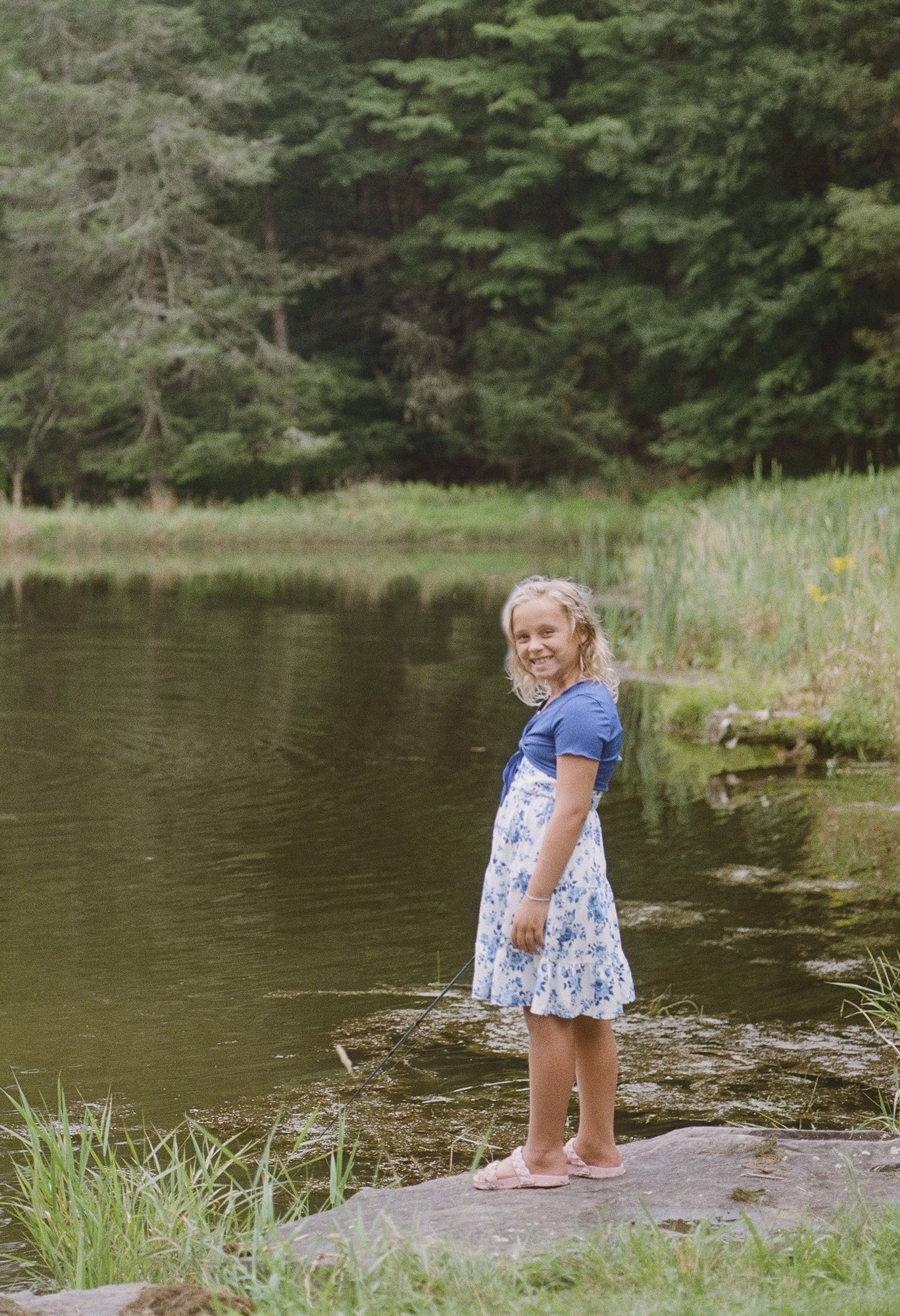 A girl with blonde hair wearing a blue top, floral skirt, and pink sandals, stands by the edge of a river or lake, holding a fishing rod and smiling. The background has tall evergreen trees and grassy areas.