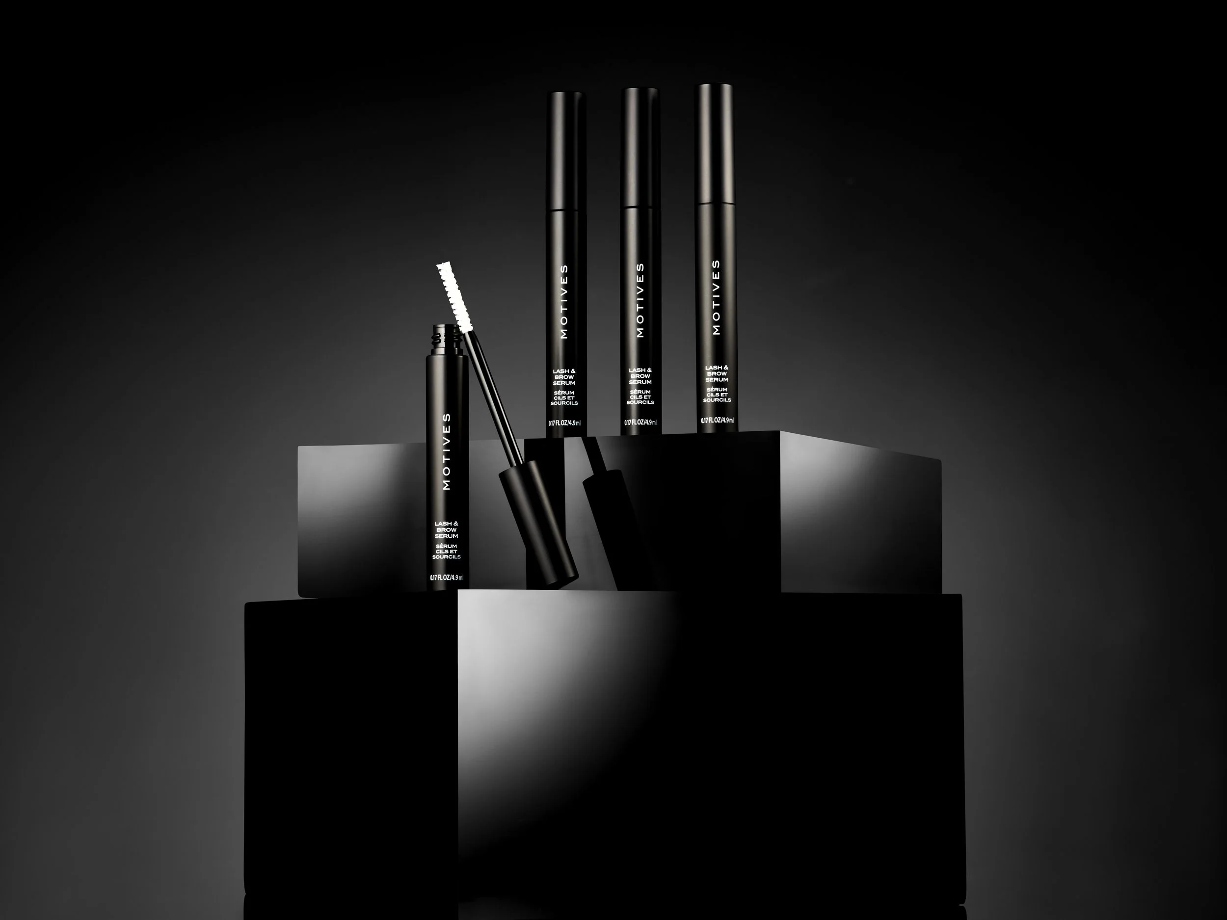 Black and white photo of MOTIVES lash and brow serum packaging, with one serum tube opened. The tubes are displayed on black platforms against a dark background.