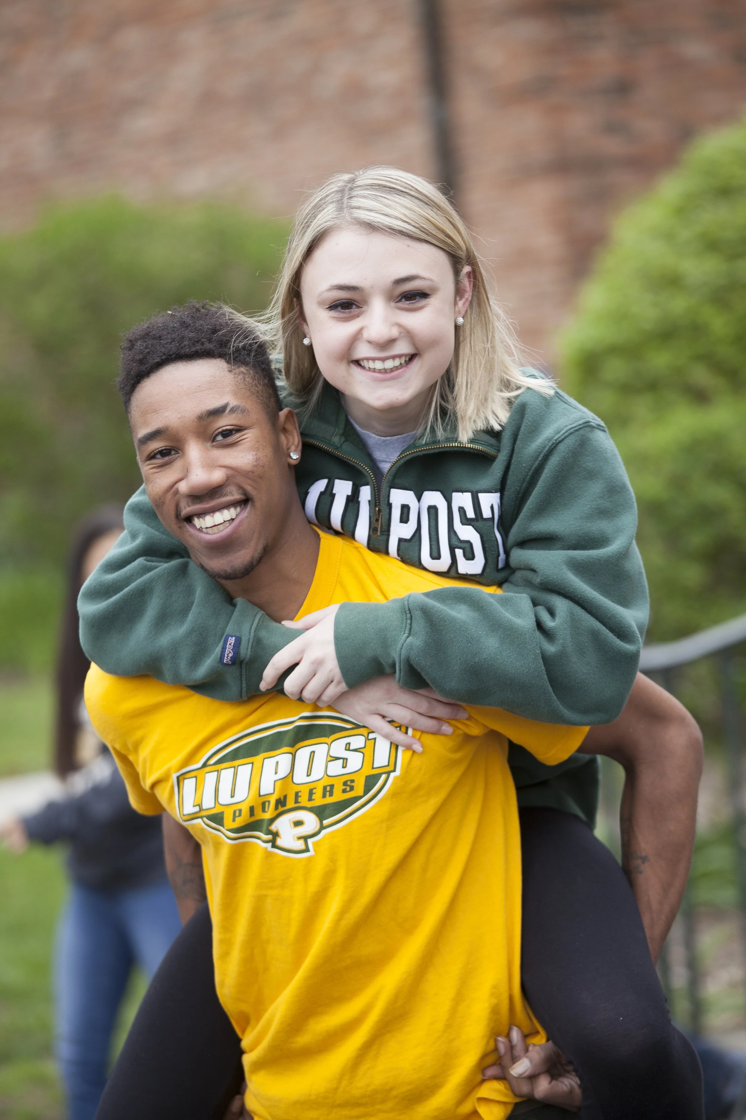 A young man in a yellow LIU Post Pioneers T-shirt is carrying a smiling young woman on his back, who is also smiling and wearing a green hoodie with LIU Post Pioneers written on it. They are outdoors with blurred greenery and a brick wall in the back