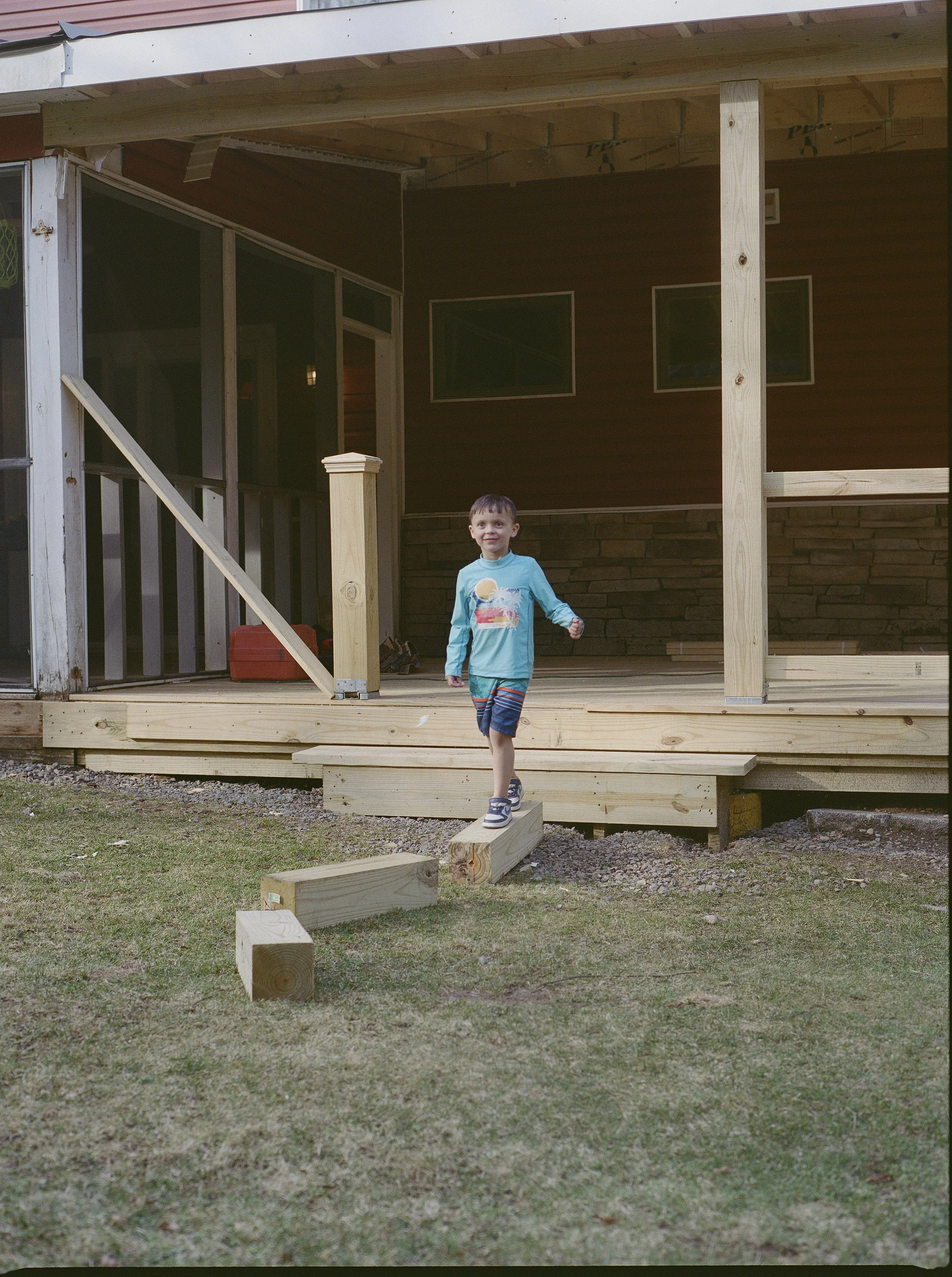 A young boy in a blue shirt with a rainbow and cloud design, and striped shorts, standing on a small wooden block in the backyard of a house that is under construction or renovation. The house has a wooden porch and parts of the structure are unfinis
