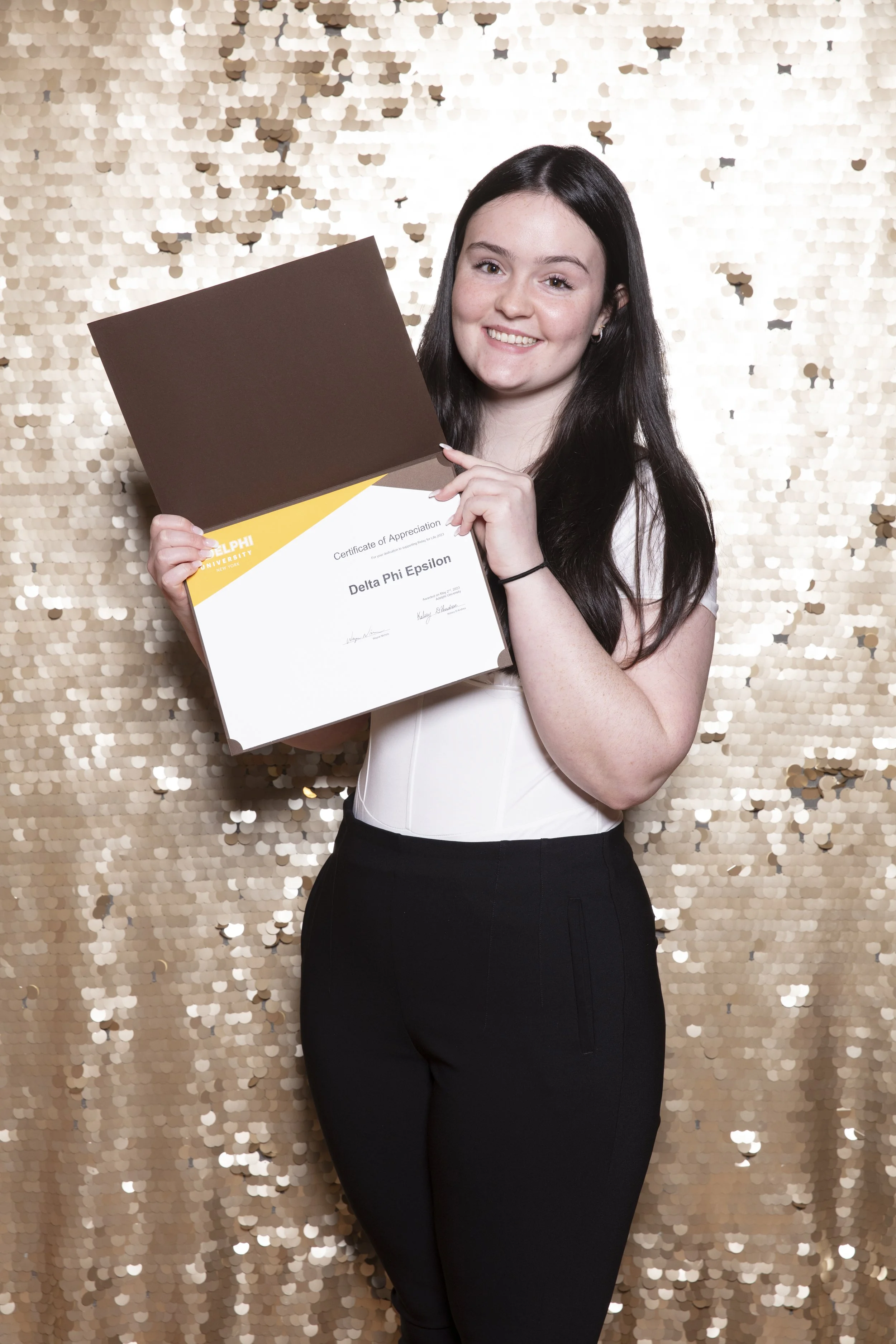A young woman with long dark hair smiling and holding a certificate of appreciation from Delta Phi Epsilon in front of a gold sequin backdrop.