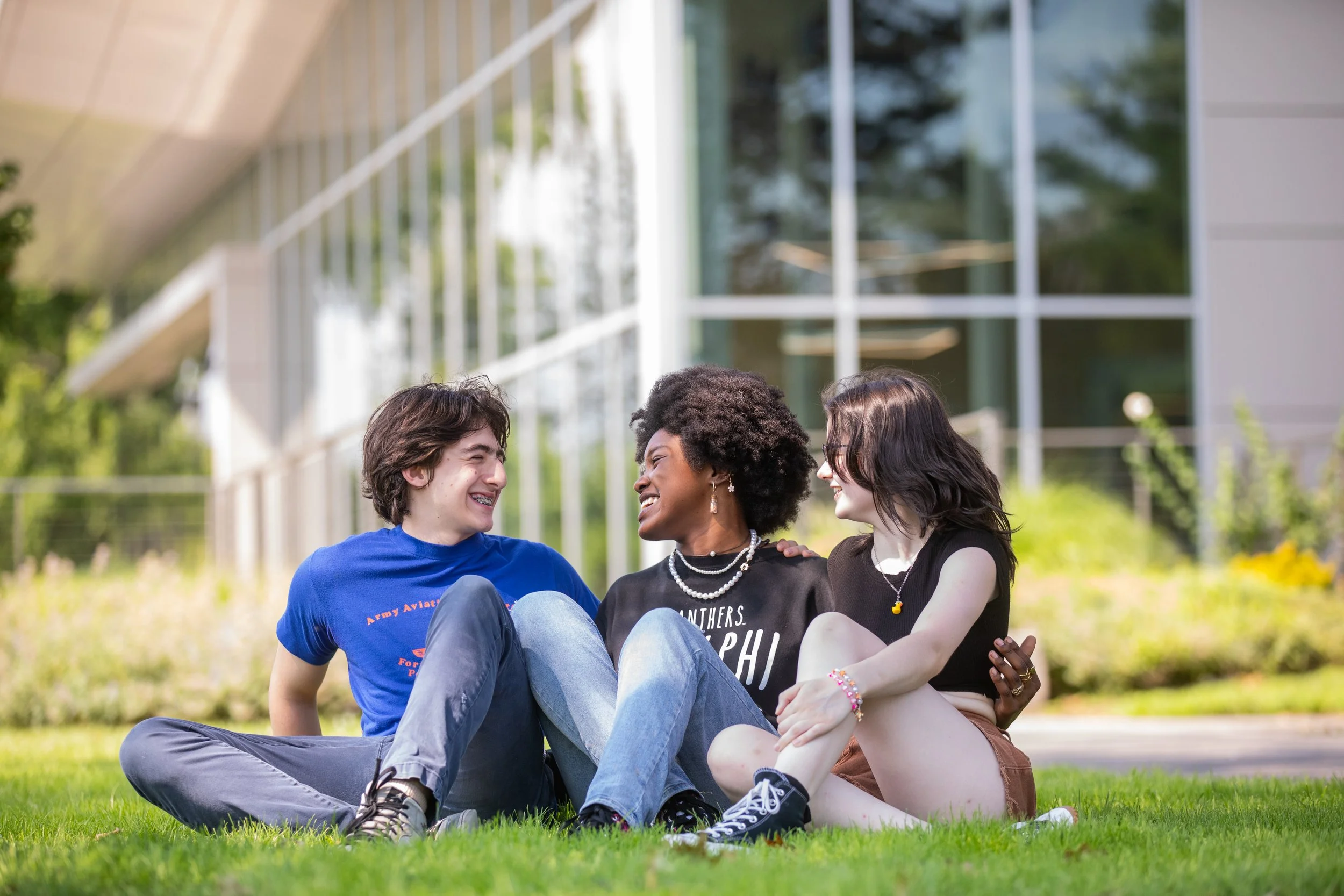 Three friends sitting on grass outside, smiling and laughing together.