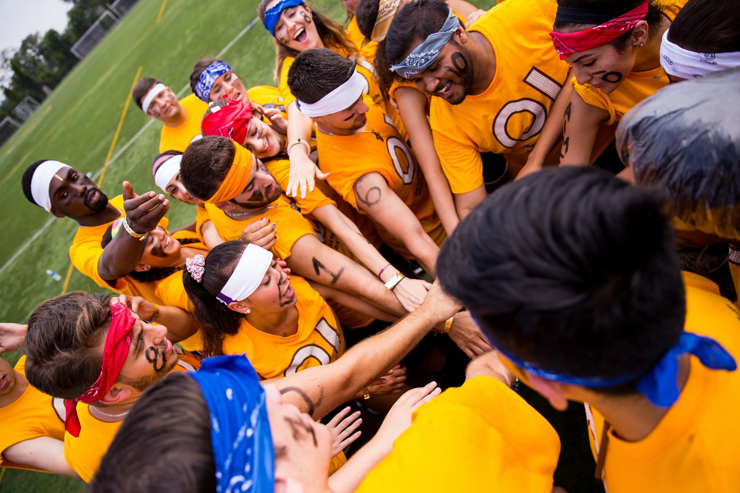 A group of young people wearing yellow T-shirts, colorful bandanas, and face paint, participating in a team activity outdoors on a grassy field. They are huddled together with their hands stacked in the center, smiling and showing camaraderie.