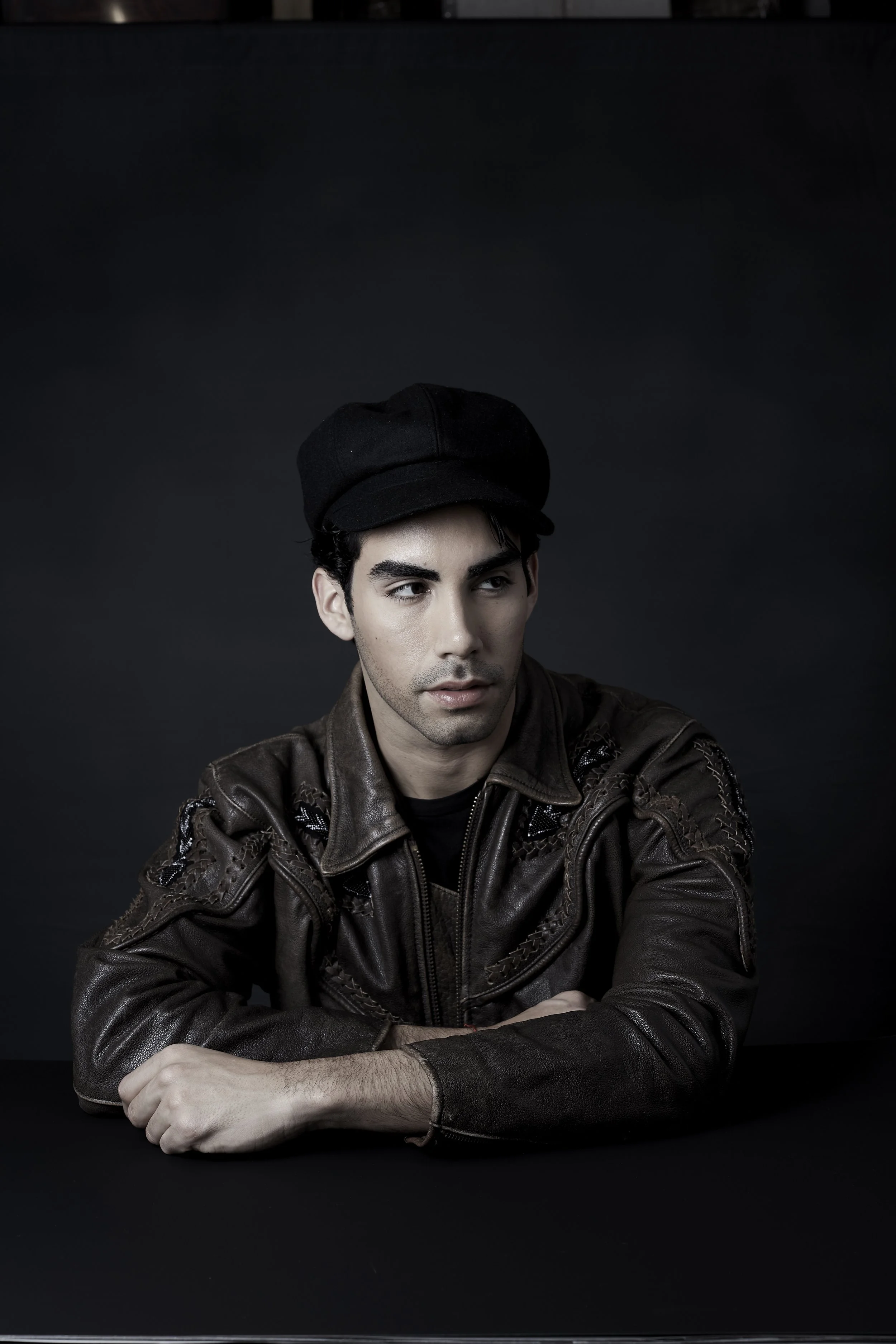 A young man with dark hair and light skin, wearing a black hat and a brown leather jacket, looks to the side against a dark background.