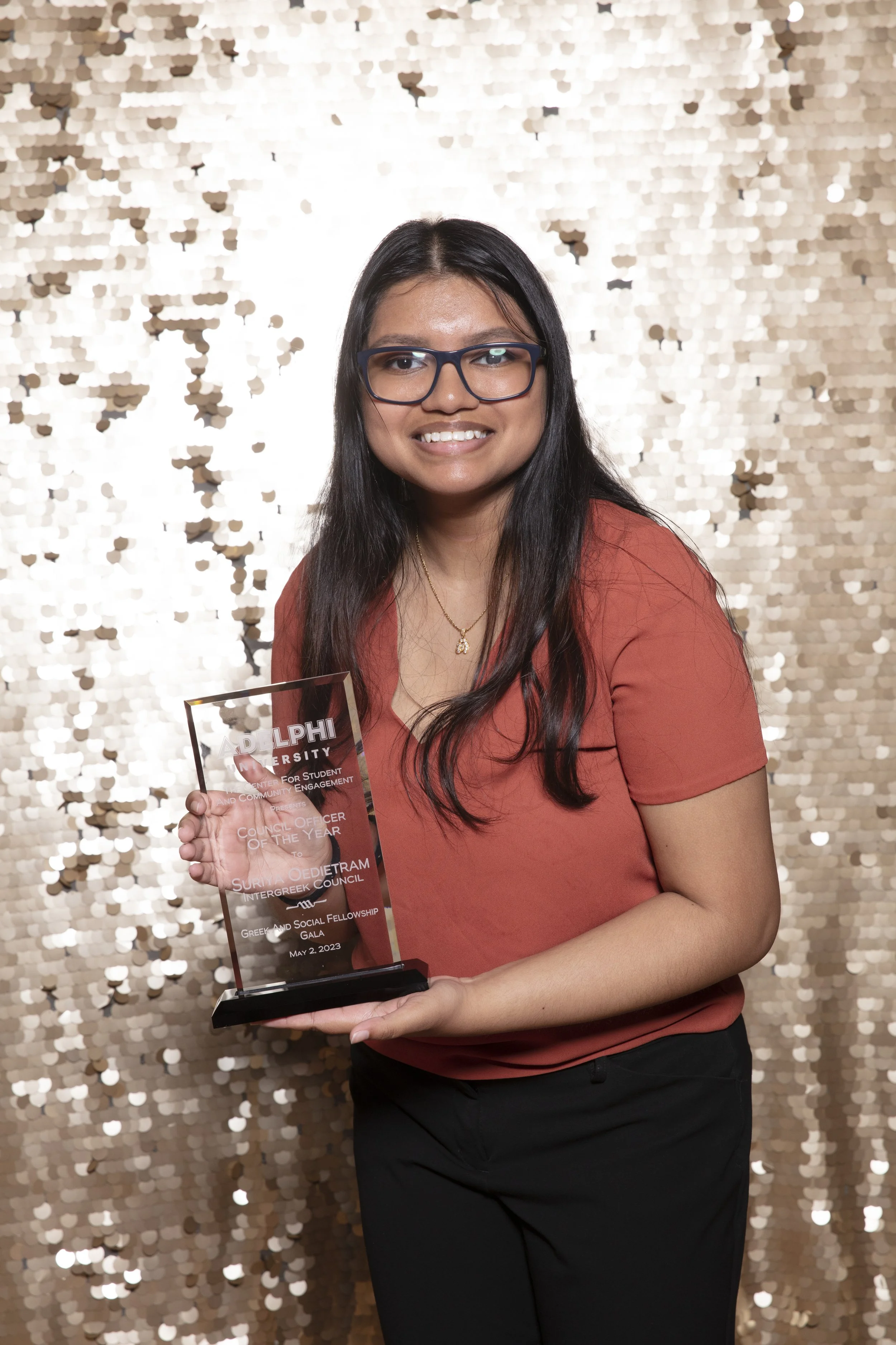 A woman with black hair, glasses, and a coral blouse holds an award at a formal event with a glittery gold backdrop.