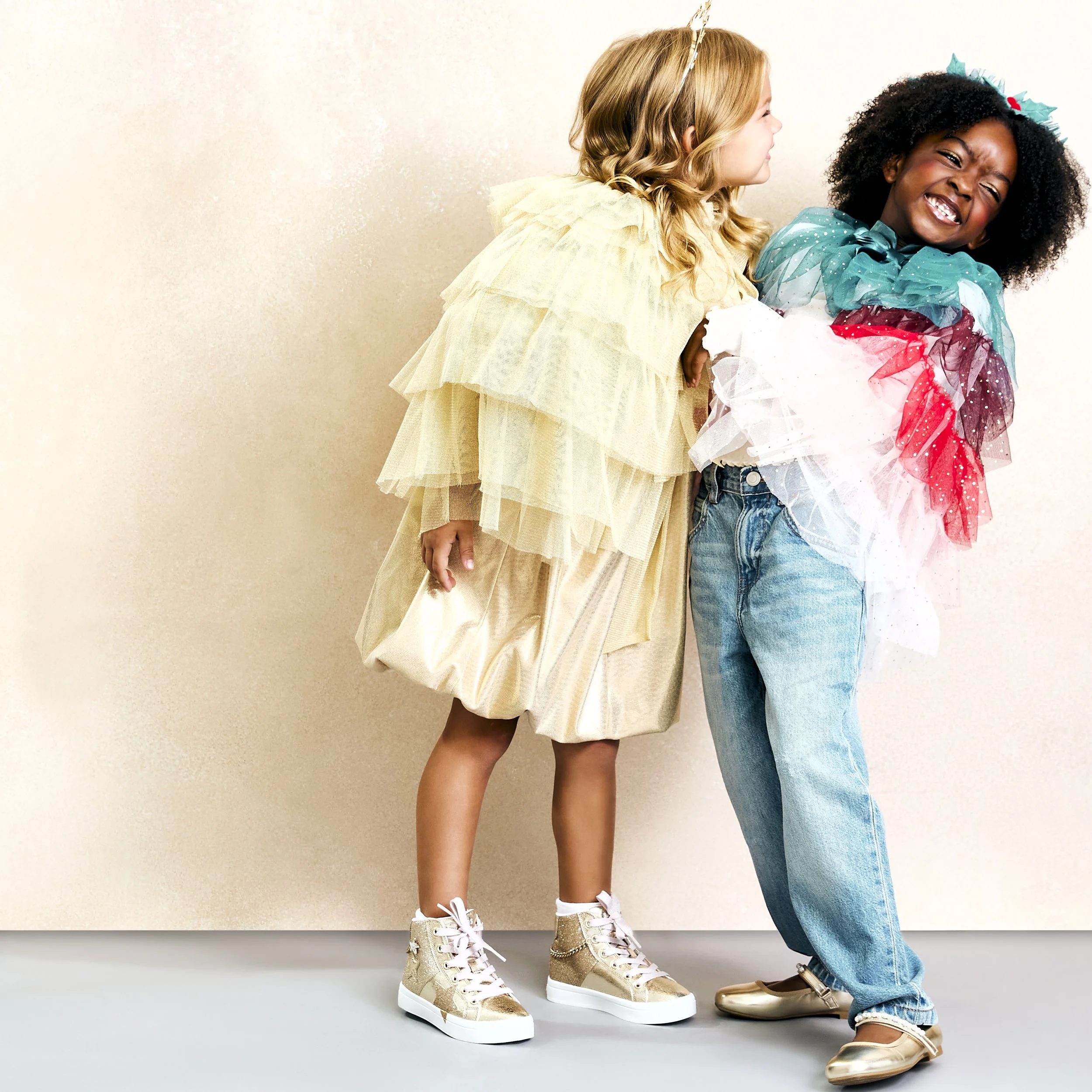 Two young girls, one in a gold dress and the other in jeans with colorful tutu layers, smiling and leaning towards each other against a plain background.