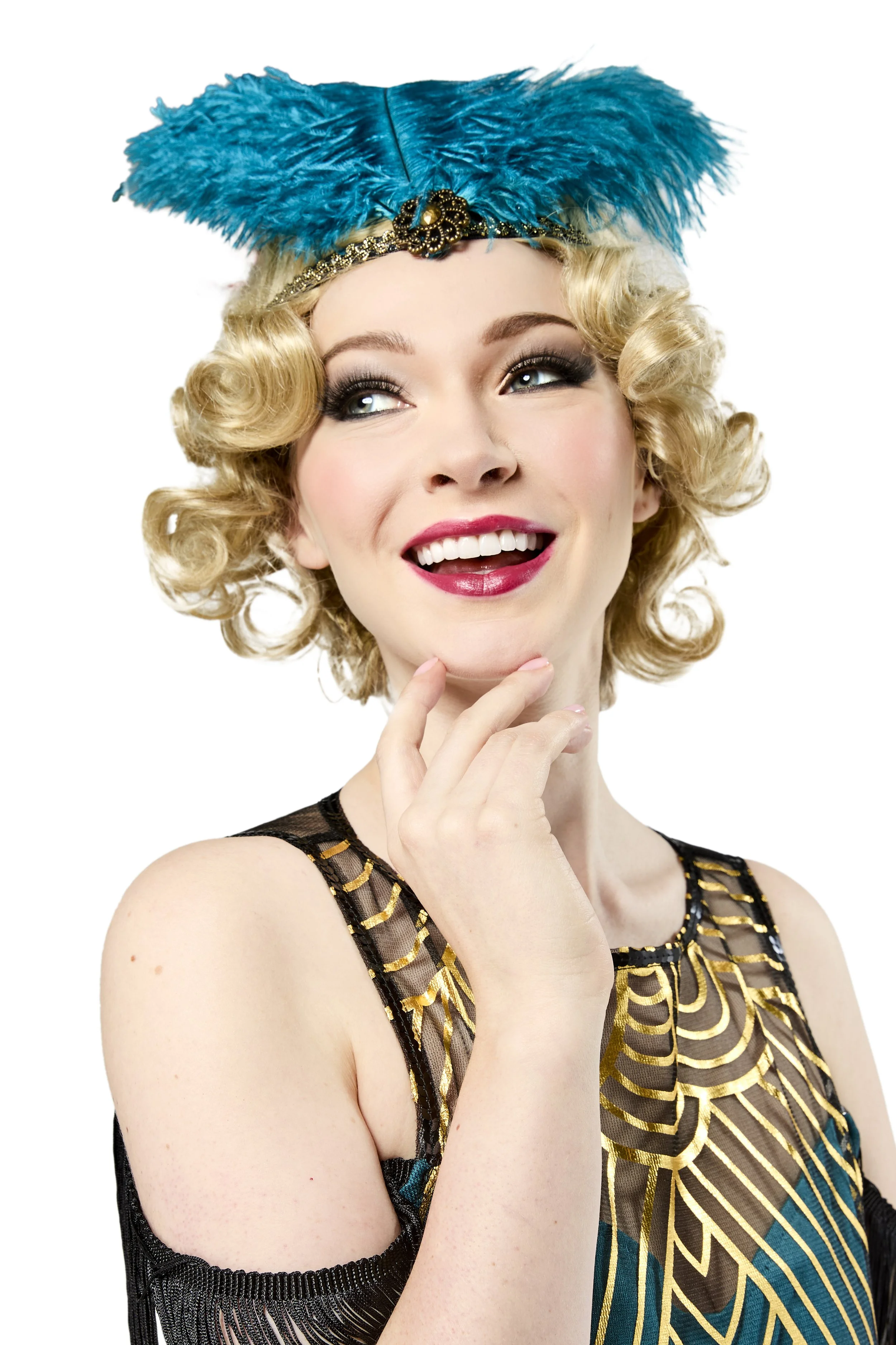 A woman with blonde curls, wearing a blue feathered headpiece and a black dress with gold and teal accents, smiling and touching her chin.