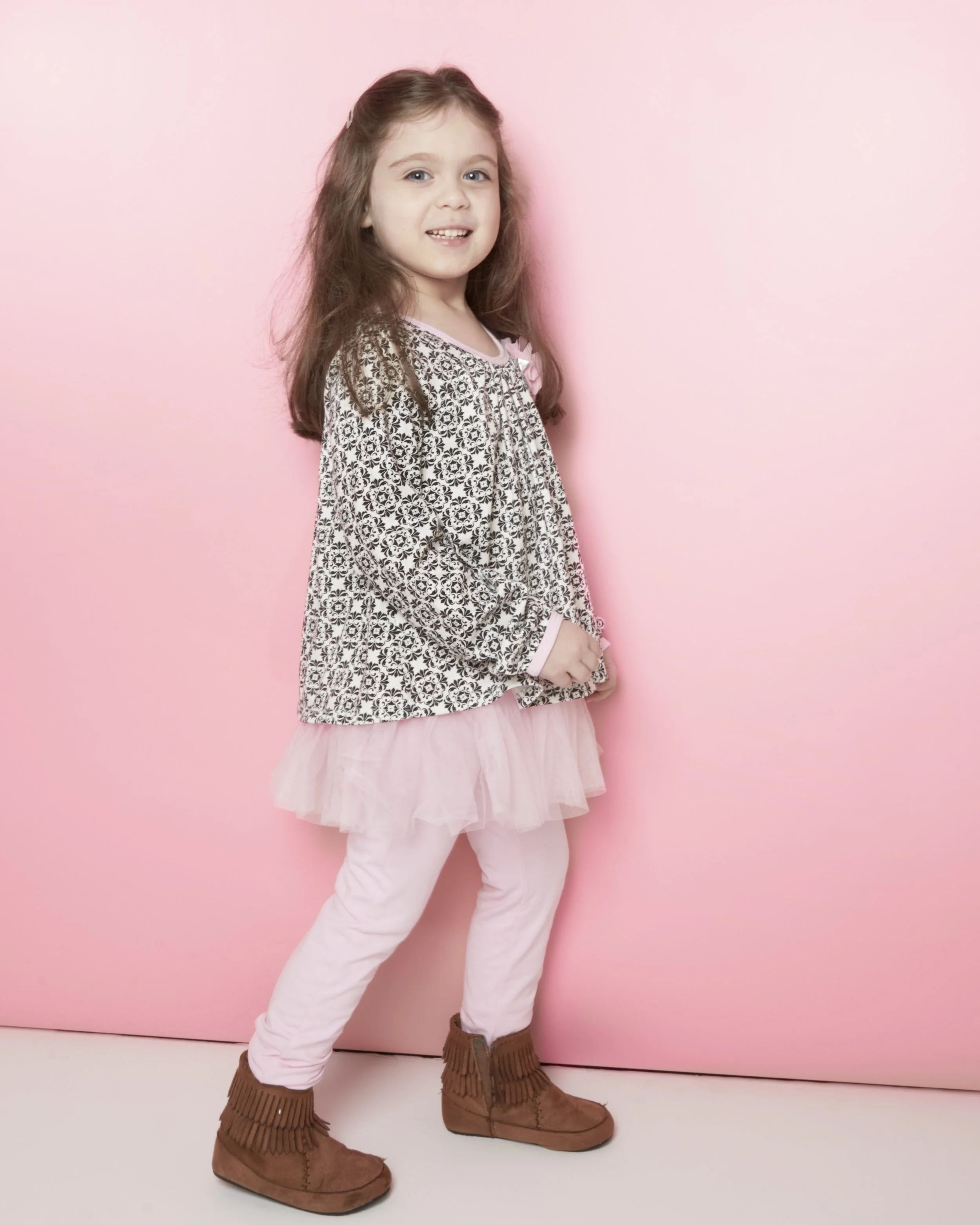 A young girl with long brown hair is standing against a pink background. She is wearing a patterned blouse, a pink tutu skirt, white pants, and brown fringe boots.