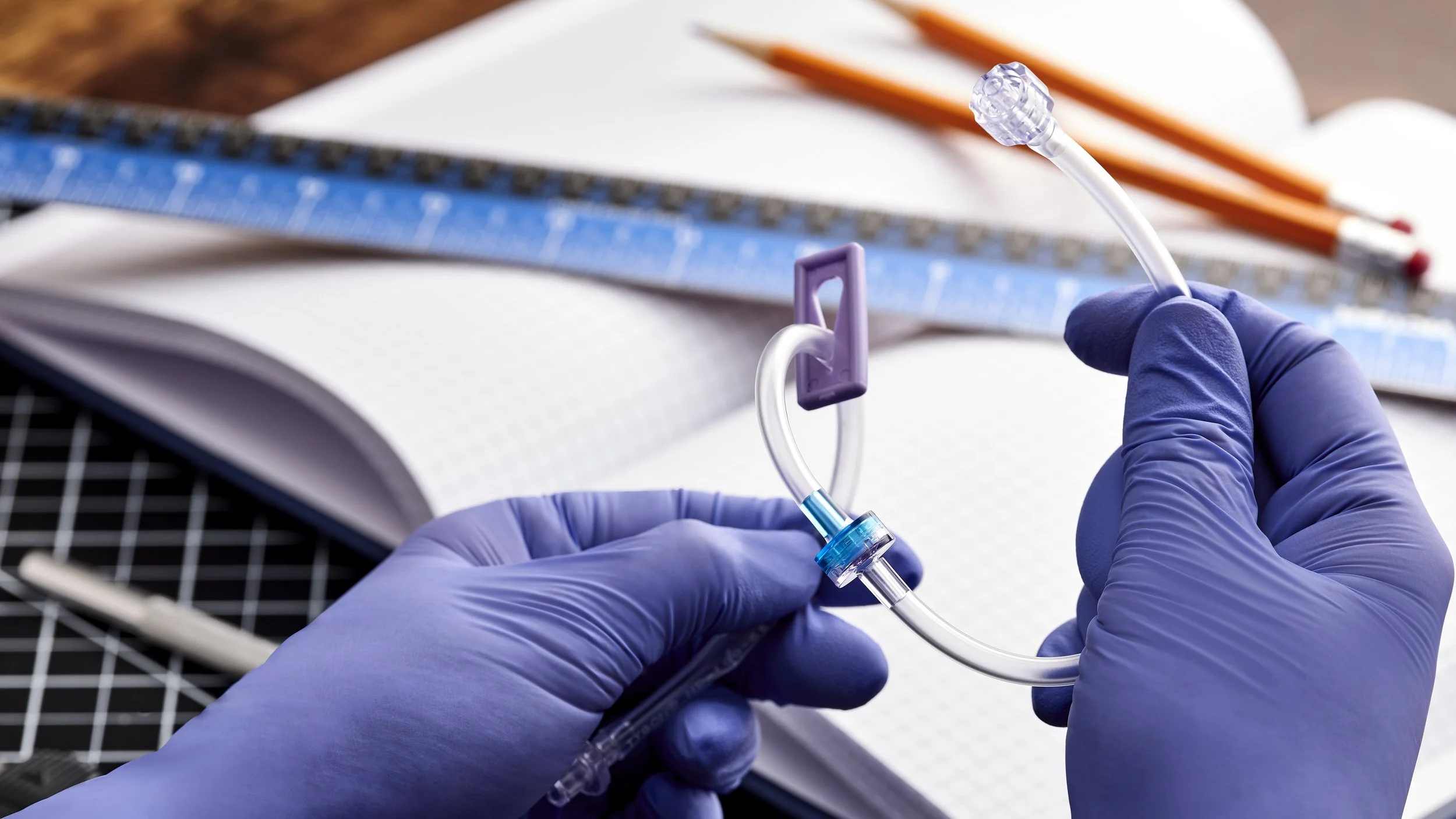 Hands wearing blue gloves holding a medical IV line with a needle and a blue connector, with a clipboard, pencils, and medical supplies in the background.