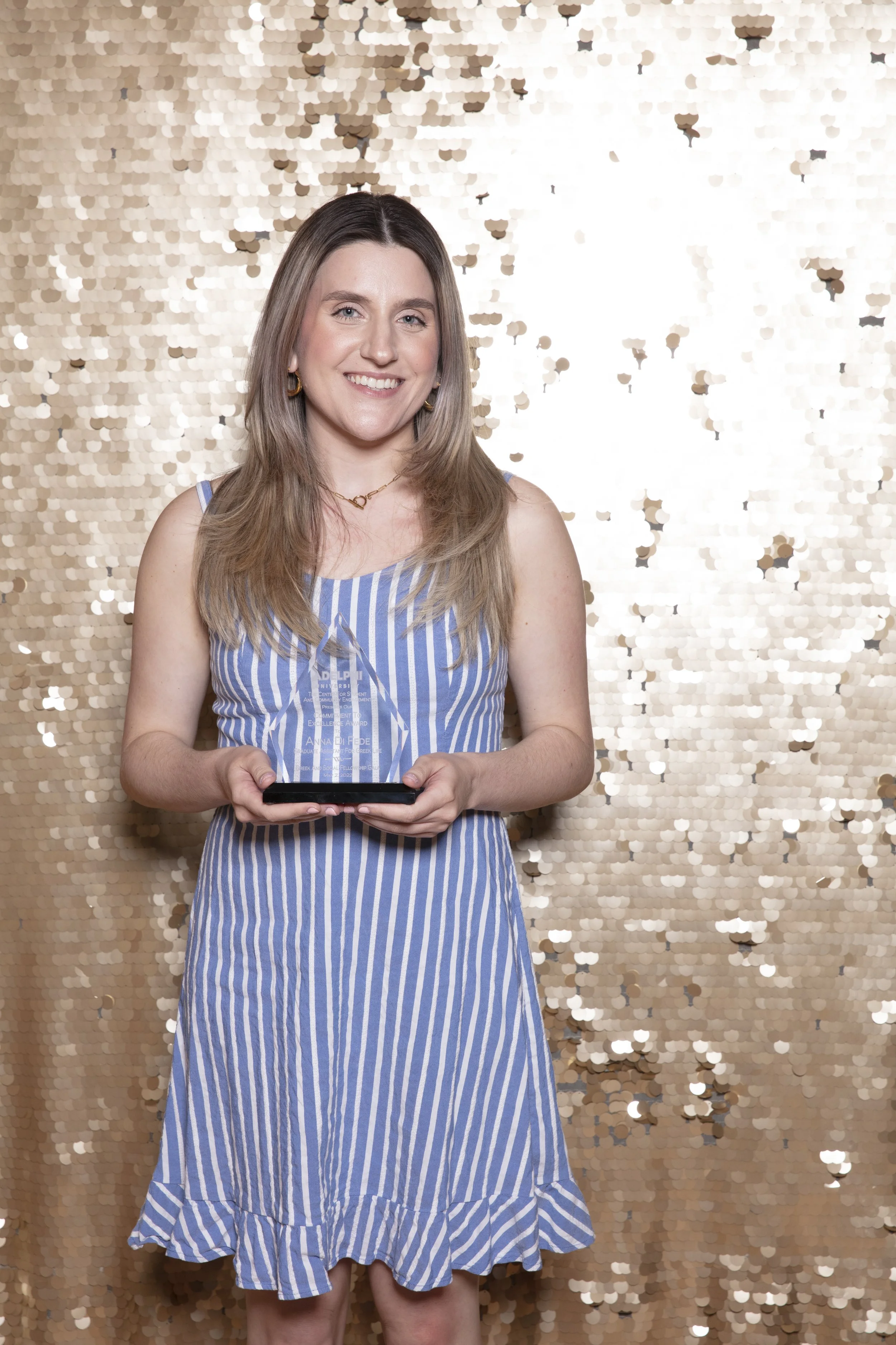 A woman with long light brown hair is holding a clear award trophy, standing in front of a gold sequin backdrop, smiling at the camera.